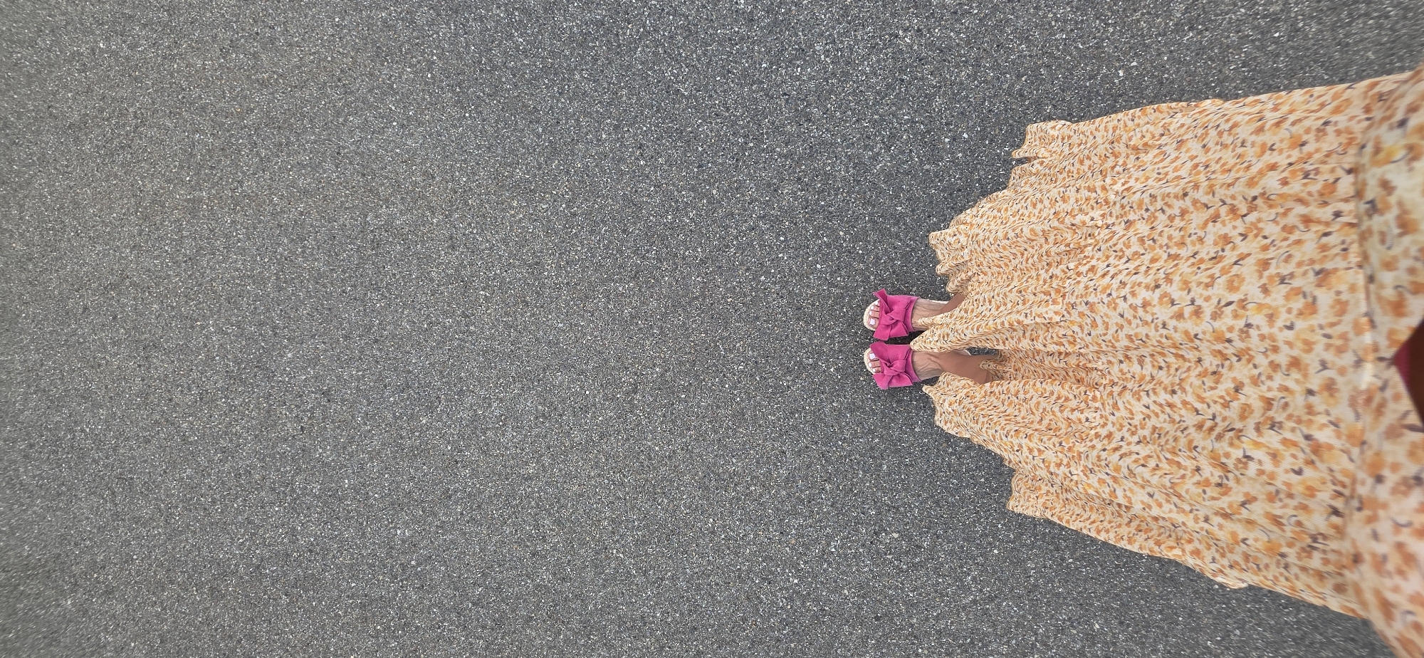A top-down perspective of a person's feet and lower legs wearing a flowing, light-colored, floral-patterned maxi dress and bright pink bow sandals. The individual is standing on an asphalt surface. The image provides no information about the city of Lieli, Switzerland.