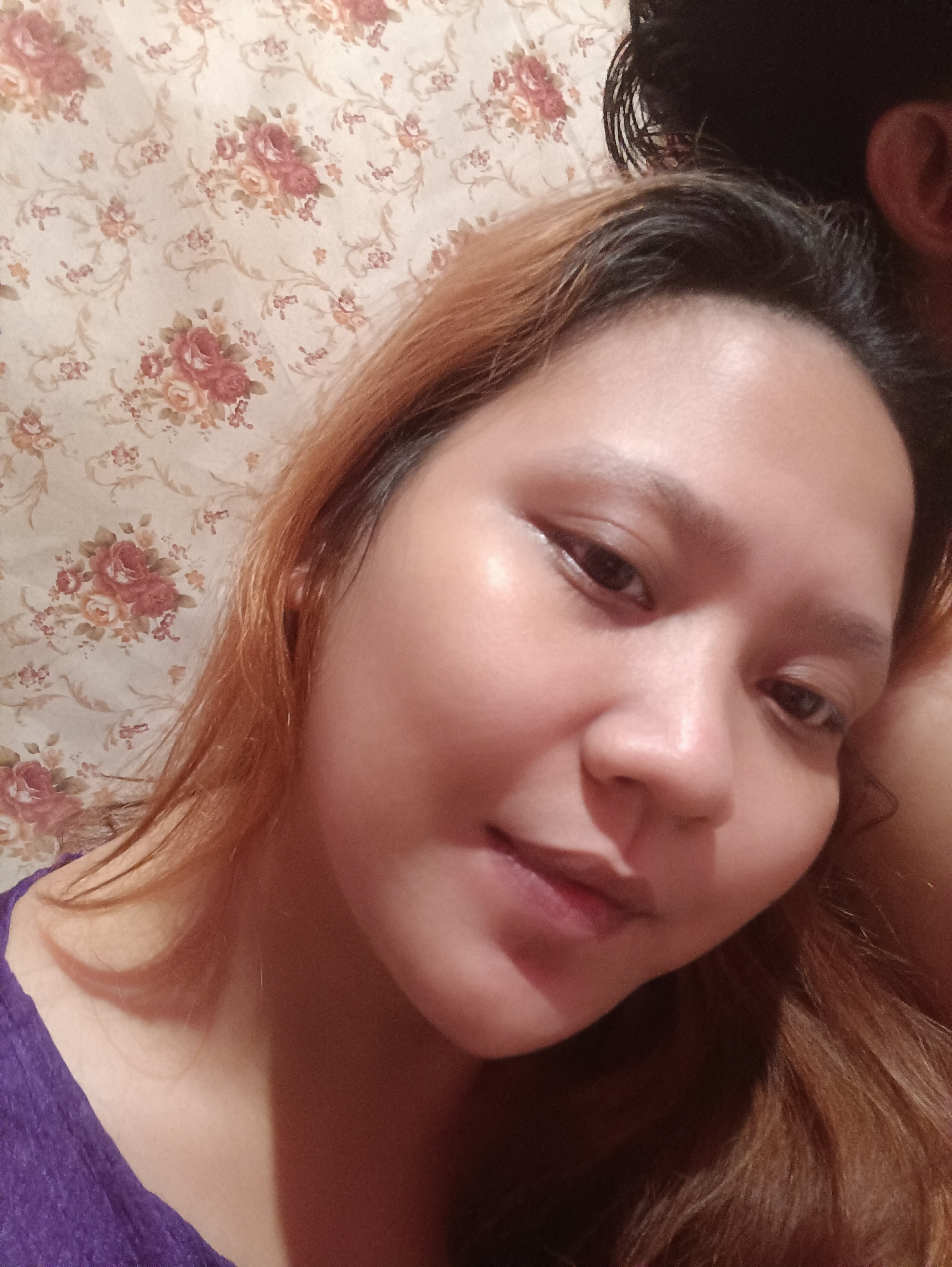 A medium shot selfie features a woman with shoulder-length, auburn hair, and a purple top. She is positioned in front of a cream-colored wallpaper with a floral pattern of pink and red roses. The woman's face is angled towards the camera, and her right ear is visible. Part of another person's dark hair and ear are visible in the upper right portion of the image, suggesting they are close to the subject. The lighting appears to be warm and slightly diffused. The scene is likely indoors, possibly in a residential setting in Depok, Indonesia, given the context of the request.