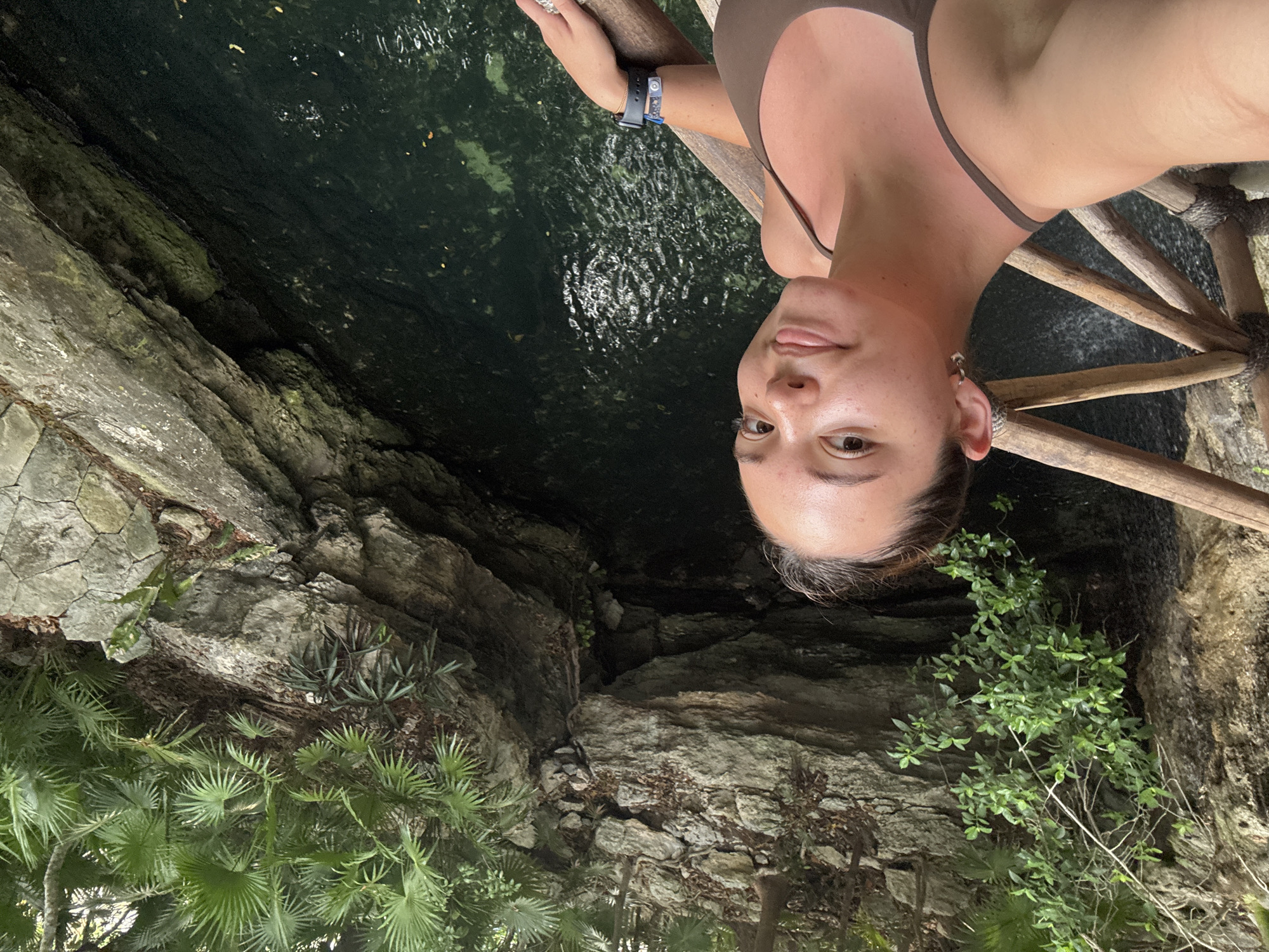 A person wearing a swimsuit and a smartwatch is on a wooden structure overhanging a deep pool of water in a cenote in Playa del Carmen, Mexico. The cenote is surrounded by limestone rock formations and vegetation, including palm trees. The water in the cenote is dark and reflective.