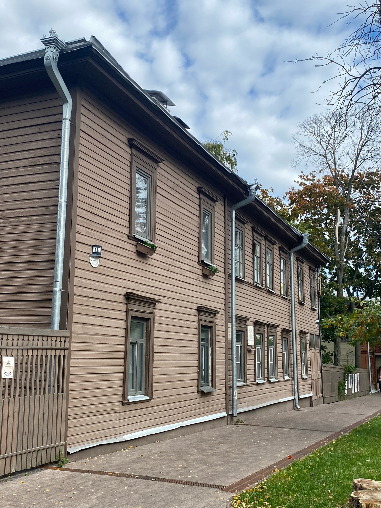 The image shows the exterior of a two-story wooden building in Nizhny Novgorod, Russia, with a brown wood-paneled facade. The building has several windows with dark trim, some of which have small planter boxes beneath them. A silver downspout runs vertically along the side of the building, attached to the gutter system. A wooden fence with vertical slats is visible on the left side of the image, and a paved walkway runs along the front of the building. The sky is partly cloudy, and trees with autumn foliage are visible in the background. A sign with the number "11a" is mounted on the building near a downspout.