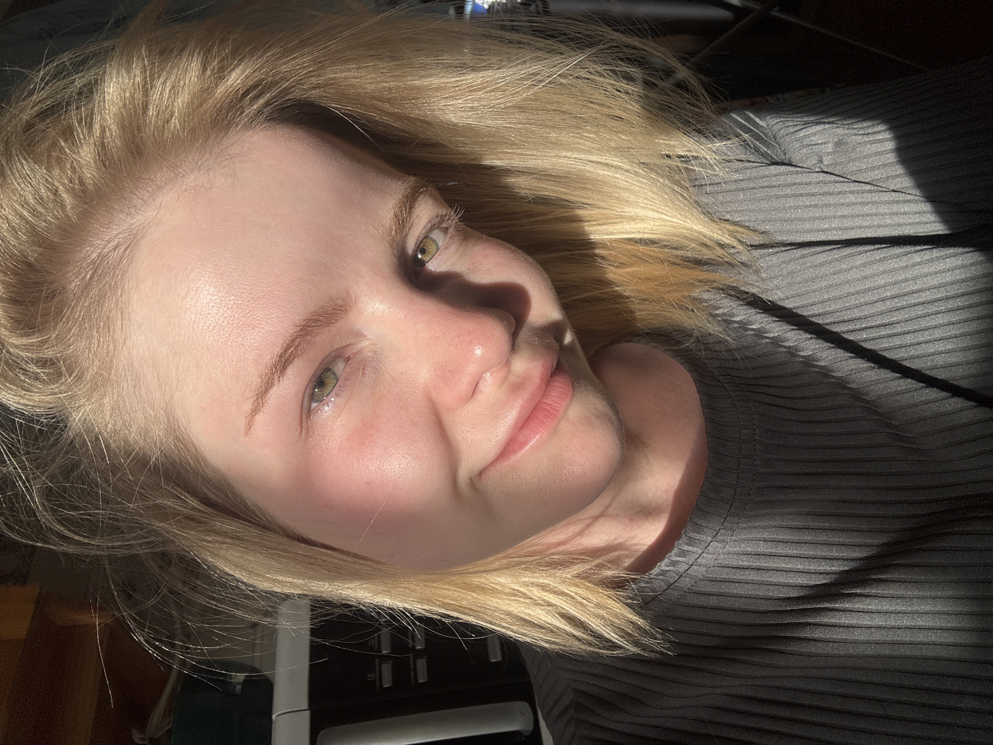 A close-up, waist-up photograph shows a young woman with blonde hair and green eyes looking towards the camera. The lighting suggests it is daytime, with sunlight casting shadows across her face and body. She is wearing a ribbed gray shirt. The background is out of focus but appears to be an indoor setting with some electronic equipment visible on the left side. The image was taken in Saint Petersburg, Russia.