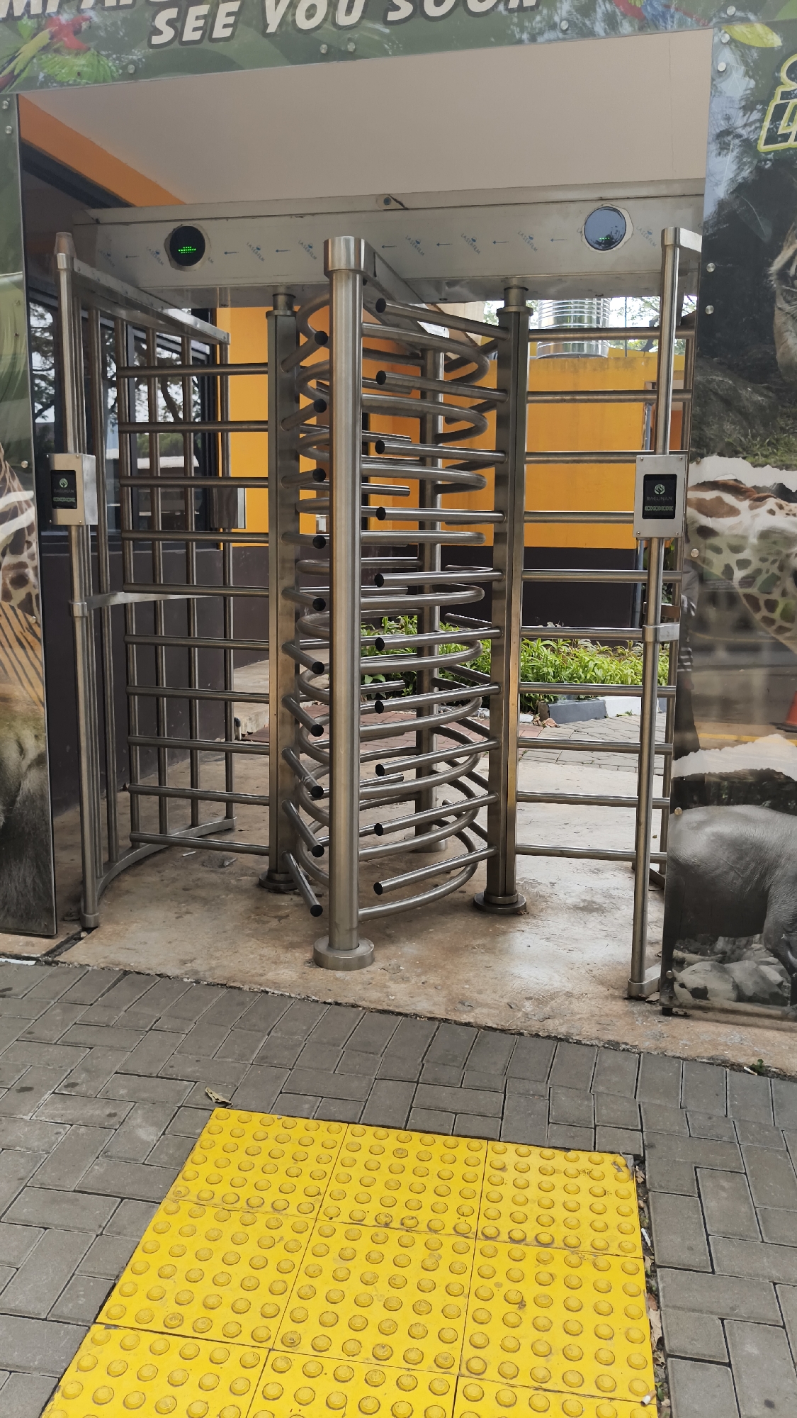 This image depicts a turnstile entrance at a facility, likely a zoo or park, located in South Jakarta, Indonesia. The turnstiles are made of brushed metal and are designed to control pedestrian flow. On the left side, there is a ticket scanner or access control device mounted on a metal post. The background features a bright orange wall and some greenery, suggesting an outdoor setting. In the foreground, there is a yellow tactile paving area, commonly used to assist visually impaired pedestrians. Large photographic murals of animals, including a giraffe and an elephant, are visible on the sides of the entrance, further supporting the idea of a zoo or wildlife park. The ground is paved with concrete and interlocking bricks. The overall scene is well-lit, indicating it was taken during daytime. There are no people visible in the immediate vicinity of the turnstiles.