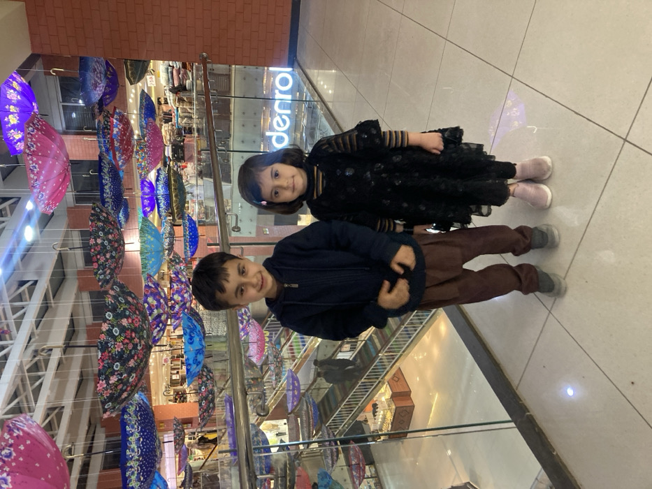 The image depicts the interior of Denrol Hyper Mall in Quetta City Tehsil, Pakistan. In the foreground, two children stand facing the camera. A male child, approximately 5-7 years old, is on the left, wearing a dark blue zippered hoodie, brown loose-fitting trousers (shalwar), and grey shoes; he exhibits a slight smile. A female child, approximately 3-5 years old, is on the right, wearing a black long-sleeved dress with a textured overlay and pink shoes; she has a neutral expression.
The background reveals a multi-level shopping mall environment. Dominating the upper portion of the frame is an extensive ceiling art installation comprising dozens of inverted, open umbrellas suspended at varying heights. These umbrellas display a variety of colors, including pink, blue, purple, black, and green, with diverse patterns such as floral and solid designs. Below the children, a transparent glass railing separates the foreground from lower levels of the mall, where an escalator is visible with two indistinct adult figures ascending. Retail areas, displaying merchandise and illuminated signage, are visible across the different levels. A prominent neon sign reading "denrol" is visible on a glass partition in the mid-ground, to the right of the children. The floor in the immediate foreground consists of large, light-colored square tiles, and a reddish-brown brick-patterned wall section is visible on the far right. The scene is brightly illuminated by artificial lighting.