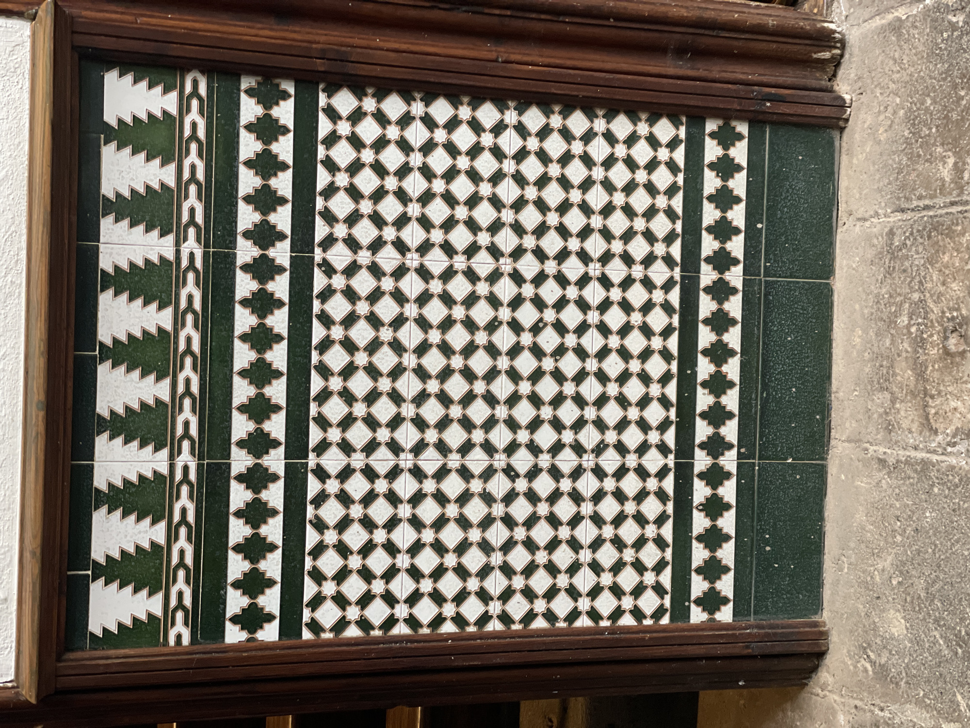 An eye-level shot captures a decorative ceramic tile panel framed by dark brown wooden molding, likely part of an interior or sheltered exterior wall in San Cristóbal De La Laguna, Spain. The tile panel displays a symmetrical, intricate design primarily in dark green and white.
The central and widest section features a repeating geometric pattern of dark green squares rotated 45 degrees, interconnected by smaller white squares and star-like shapes, creating a lattice effect. Flanking this central pattern are two narrower vertical bands, each showcasing a series of dark green trefoil or quatrefoil-like shapes outlined in white, set against a white background. The outermost vertical bands (closest to the solid dark green edges) are composed of a repeating zigzag or stepped pattern in dark green against white, resembling stylized trees or arrows. The entire tile arrangement is subtly outlined in a lighter, off-white or beige tone.
To the left of the wooden frame, a plain white wall is visible. To the right, a rough-textured, light gray or beige stone wall or pillar suggests an older architectural structure. The tiles exhibit a slightly glossy finish with minor imperfections, while the wooden frame shows its natural grain. The lighting is soft and even. No people or visible text are present in the image.