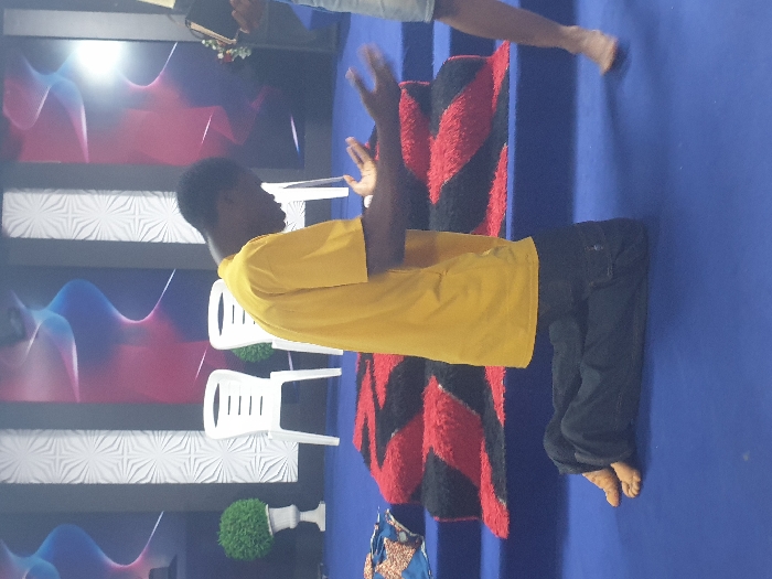 A man in a yellow shirt and dark pants stands barefoot on a blue surface. He gestures with his hands. Behind him, a red and black rug partially covers the blue floor. To the left, three white plastic chairs are visible, set against a backdrop featuring white patterned panels and screens with blue and red wave-like designs. A decorative green bush sits near the bottom left. Another person's legs and bare feet are visible in the upper right corner.