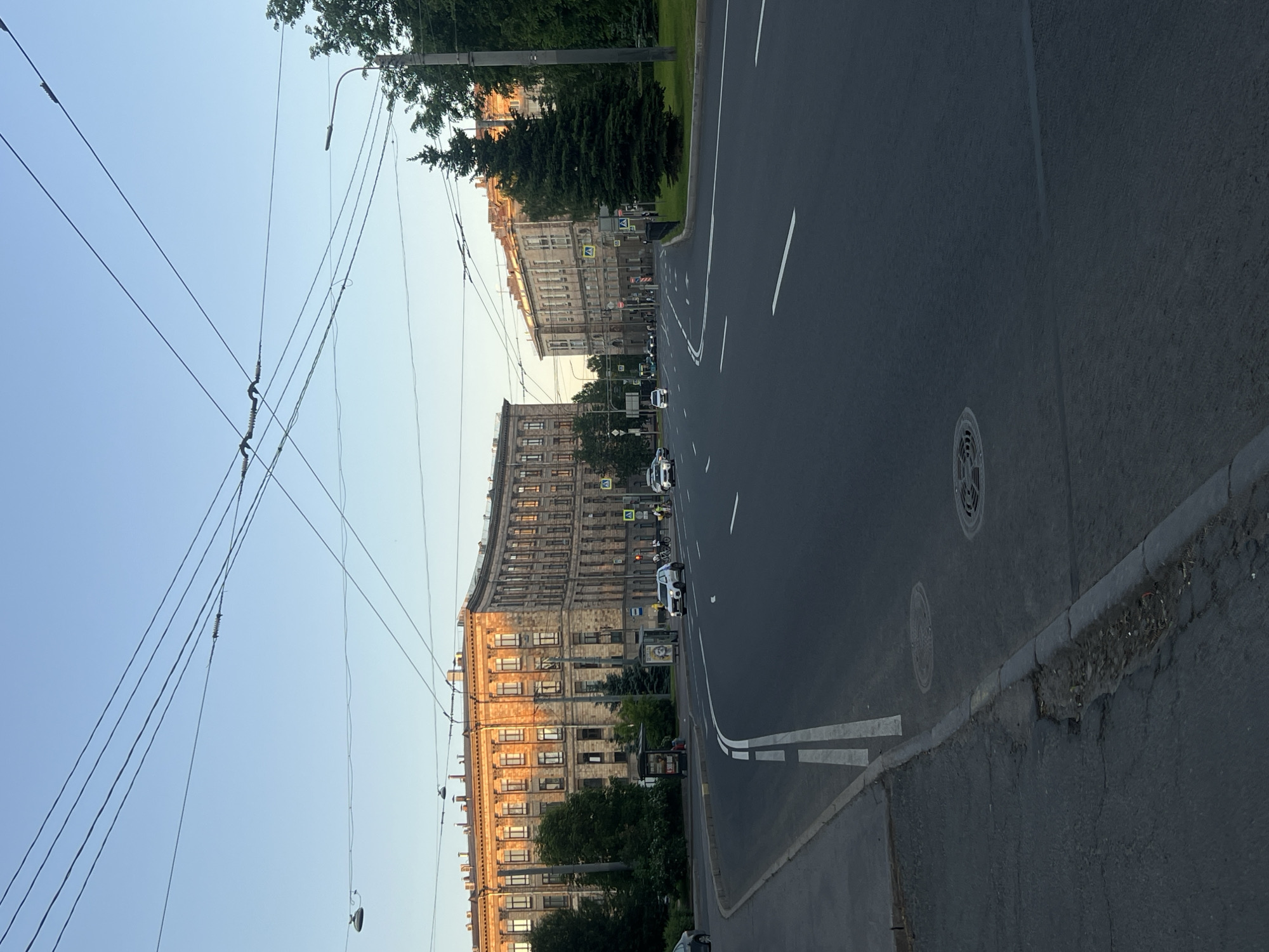 This image depicts a wide-angle street view of Ploshchad Muzhestva (Courage Square) in Saint Petersburg, Russia, under clear daylight conditions. The scene is illuminated by the low angle of the sun, casting warm light on the building facades on the left.
The upper third of the image shows a clear, light blue sky traversed by numerous black overhead electrical wires, characteristic of a trolleybus or tram system. These wires form a complex, intersecting network across the frame. A single, dark street light fixture is visible on the far left, hanging below the wires.
The left side of the image features a large, multi-story building with a distinctive curved corner, constructed from a light brown or yellowish stone facade. Its numerous rectangular windows are uniformly arranged and reflect the golden sunlight. Below the building, green deciduous trees and two dark green coniferous trees are visible, along with two bus stop shelters. The closest bus stop has an advertisement on its back panel.
The central and lower portions of the image are dominated by a wide, dark asphalt road. White solid and dashed lines delineate multiple lanes, indicating traffic flow away from the viewer. On the far left, a distinct double solid white line marks a turning lane or shoulder. Several circular metal manhole covers are embedded in the road surface in the foreground and mid-ground. The asphalt curb on the far left shows some signs of wear and minor damage.
Vehicles are sparsely distributed on the road. A white SUV is visible in the left lanes, moving away from the viewer. Further down the road, additional white cars are visible on both sides of the roadway. Several individuals on bicycles and pedestrians are discernible in the distance near crosswalks.
The right side of the image features a dense line of dark green deciduous trees and several prominent, dark green coniferous trees, separating the road from another, similar multi-story building partially visible in the distance. A tall, black street light pole with a horizontal arm is positioned on the far right. Yellow diamond-shaped pedestrian crossing signs are visible across the intersection in the mid-ground. Green grass covers the median strips and verges along the road. The overall lighting suggests either late afternoon or early morning.