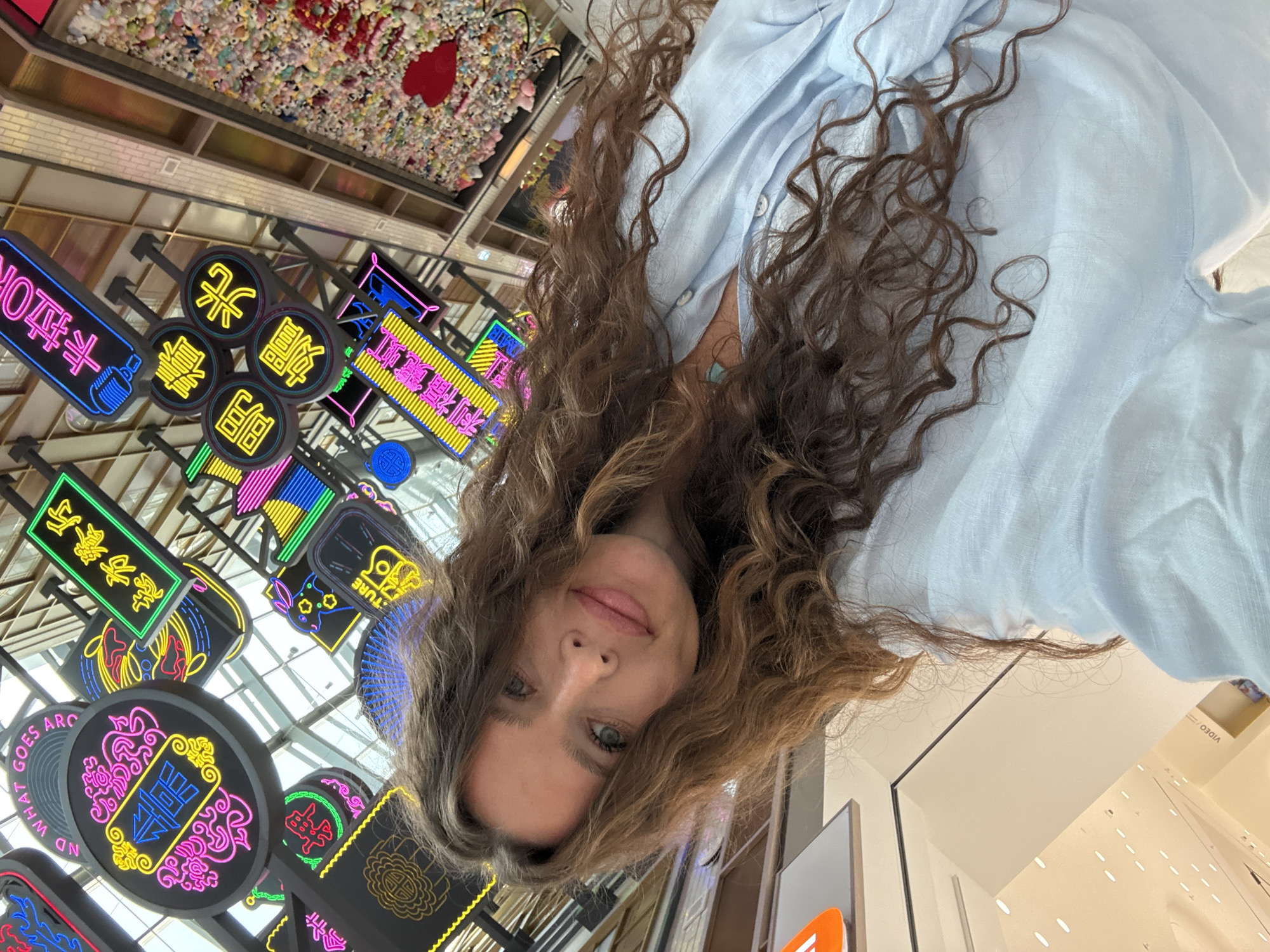 A woman with long, wavy brown hair is taking a selfie in an indoor commercial space. She is wearing a light blue button-up shirt. Behind her, a large wall is covered with numerous small stuffed toys, forming a colorful mosaic. Red heart-shaped decorations are visible on the wall. Above her head, there are many illuminated neon signs, some with Chinese characters and others with English text, such as "AND WHAT GOES ARC" and "卡拉 OK" (Kala OK). The overall environment suggests a vibrant and modern shopping mall or entertainment venue in Dubai, United Arab Emirates.