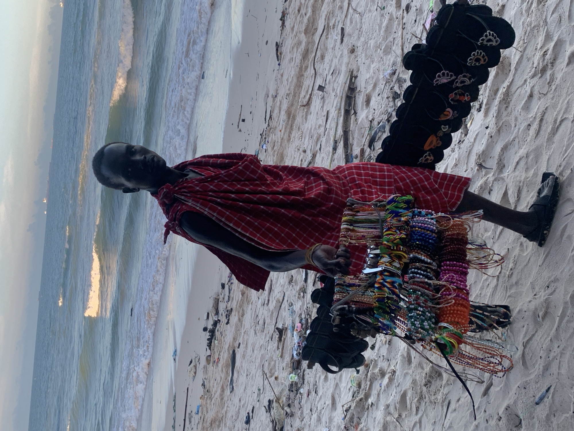 On a sandy beach in Dar es-Salaam, Tanzania, a person, likely a man, is captured in what appears to be late afternoon or early evening light. He wears a distinctive red and white plaid garment, possibly a Maasai shuka. The individual is positioned centrally, holding up a display of numerous colorful, handmade beaded bracelets and necklaces, suggesting he is a vendor.
To his right, a vertical arrangement of black flip-flops, some decorated with patterns resembling animal paw prints, is also displayed for sale. The vendor looks directly at the camera with a neutral expression.
Behind him, the calm ocean stretches to the horizon, with gentle waves breaking on the shore. A warm, golden light reflects on the water's surface, characteristic of sunset or sunrise. The sandy foreground shows scattered debris, including bits of plastic and wood. A small tag on one of the beaded bracelets is partially visible, showing the text "SPORT CLUB". The scene depicts a common sight of local craft sales along the Tanzanian coastline.