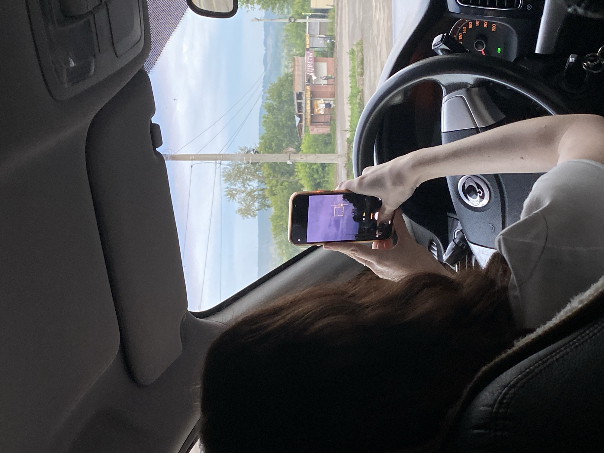 The image shows a person sitting in the driver's seat of a car, holding a smartphone and taking a picture of the landscape outside. The person's back is to the camera, and their hair is visible. The car's steering wheel and dashboard are in the foreground. Outside the car, there is a street with buildings, trees, and a cloudy sky. A sign on one of the buildings reads "ЦВЕТЫ" (flowers) in Russian. This scene is consistent with the city of Votkinsk, Russia, a city known for its historical ties to composer Pyotr Ilyich Tchaikovsky and its industrial activities. The image captures a moment of everyday life, with technology intersecting with the observation of the surrounding environment. The overall atmosphere is casual and observational.