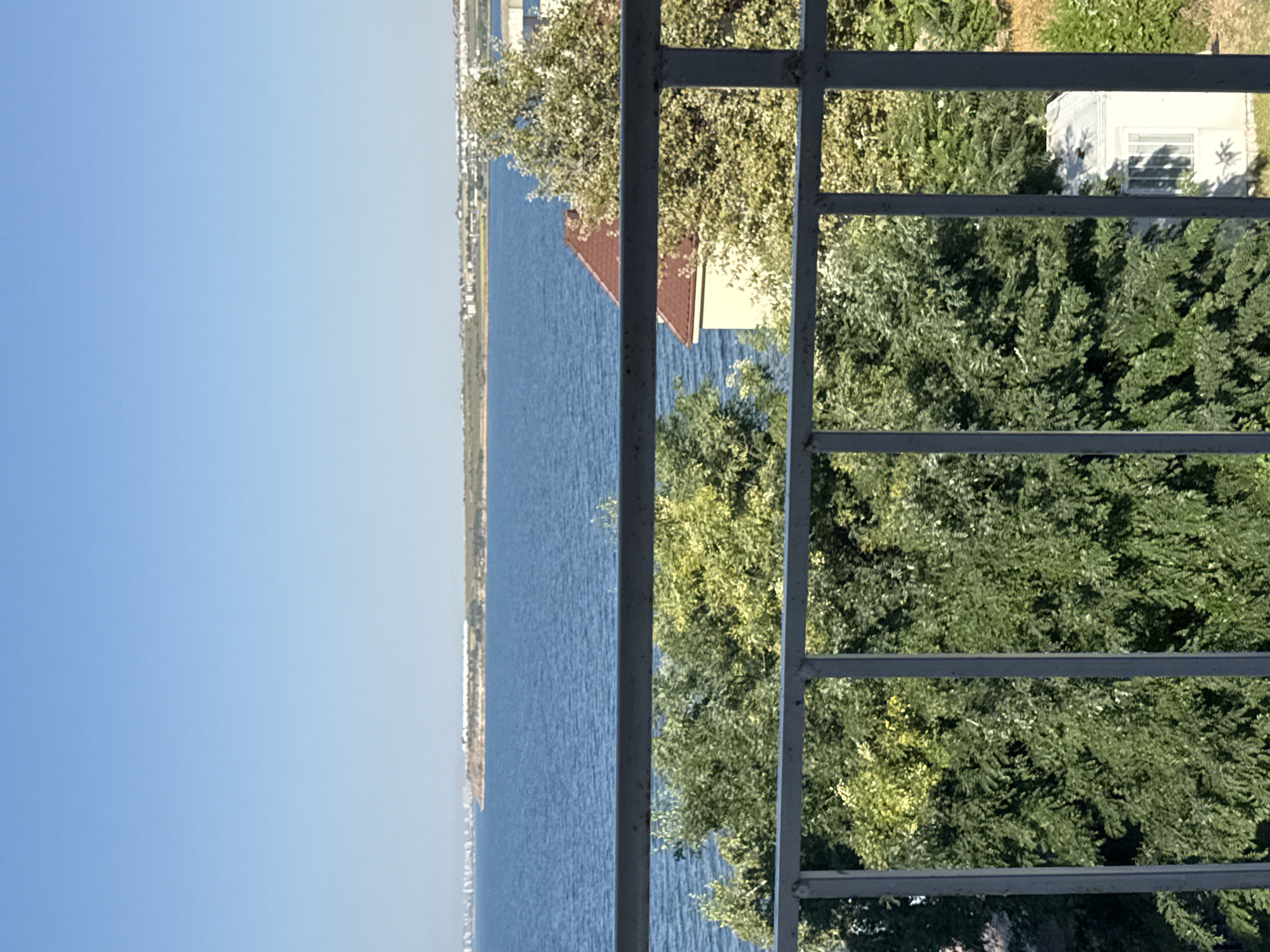 The image captures a view from a balcony overlooking a calm body of water, likely the Black Sea, with the city of Pomorie, Bulgaria, visible in the distance. The foreground is dominated by a grey metal railing, behind which lush green trees with some lighter-colored foliage fill the lower half of the frame. A portion of a building with a red-tiled roof and cream-colored walls is visible through the trees on the right. The water is a deep blue, stretching towards a distant, flat shoreline dotted with buildings and infrastructure. The sky above is a clear, bright blue, indicating a sunny day. The overall scene suggests a peaceful, scenic setting.