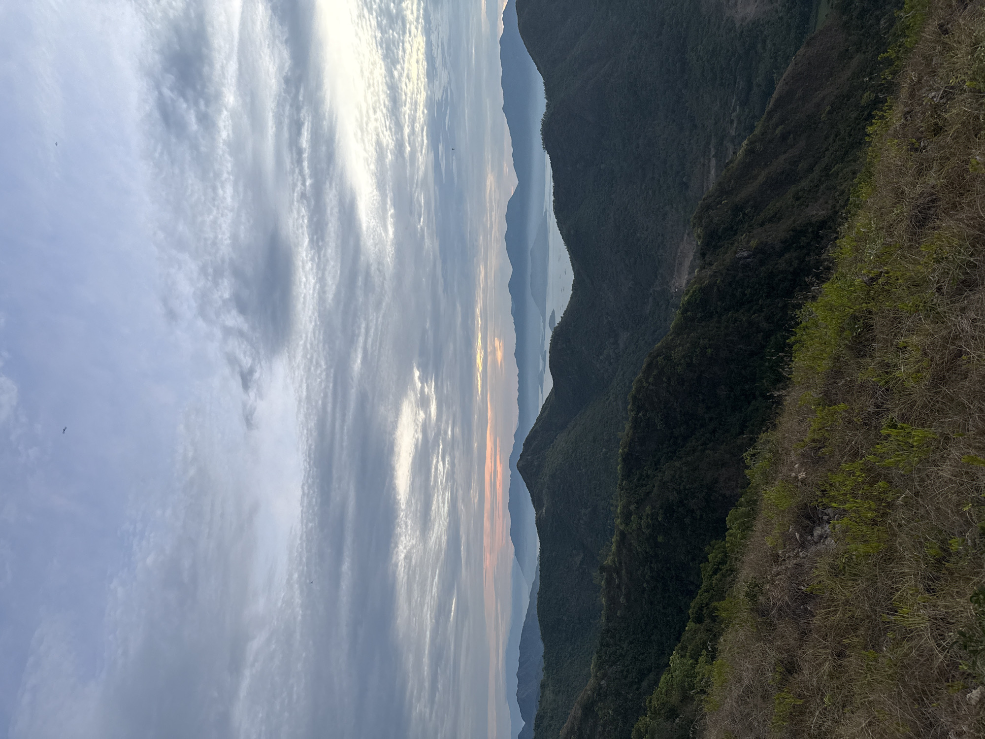 The image depicts a landscape scene with layered mountains, a body of water, and a cloudy sky. The closest mountain range is covered in lush green vegetation, while the subsequent ranges appear progressively paler and blur into the horizon, indicating distance. Between the mountains are glimpses of a body of water. The sky is filled with long, striated clouds that are illuminated by the setting sun, casting a gradient of colors from pale yellows to oranges. The location is Bac Nha Trang Ward, Vietnam.