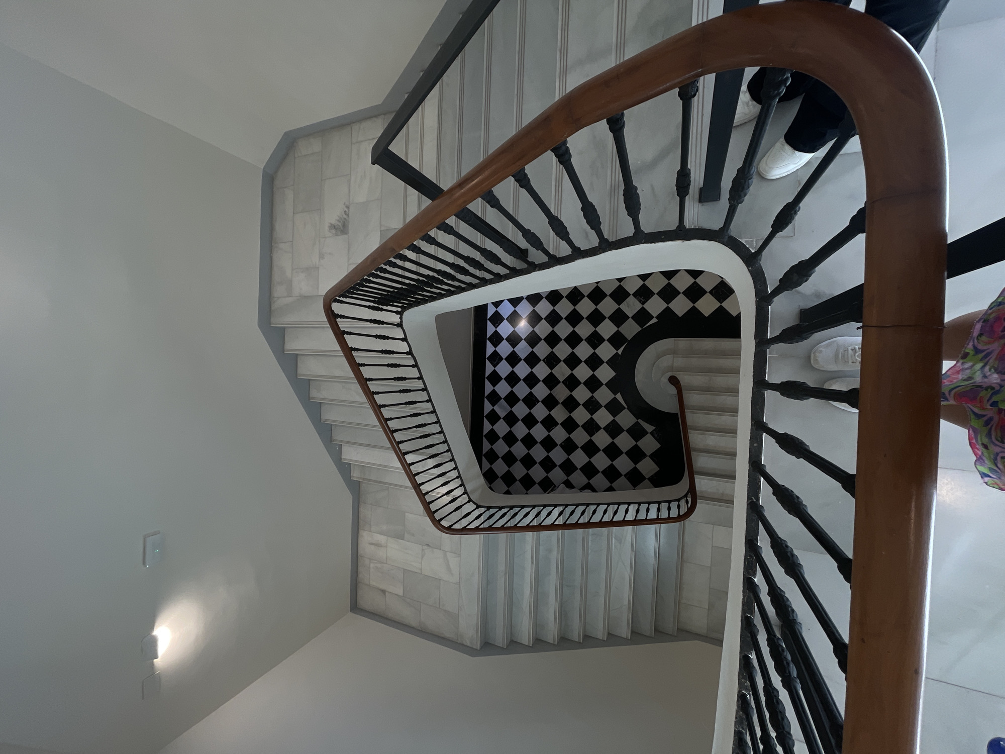 The image shows a top-down view of a staircase with a wooden handrail and black balusters. The staircase is located inside a building in València, Spain. The stairs are covered in white marble. The landing has a black and white checkered tile floor. Two individuals are partially visible in the shot. One is wearing black pants and white sneakers. The other is wearing white sneakers and a multicolored dress. There are two light switches and a light fixture on the wall to the left of the staircase.