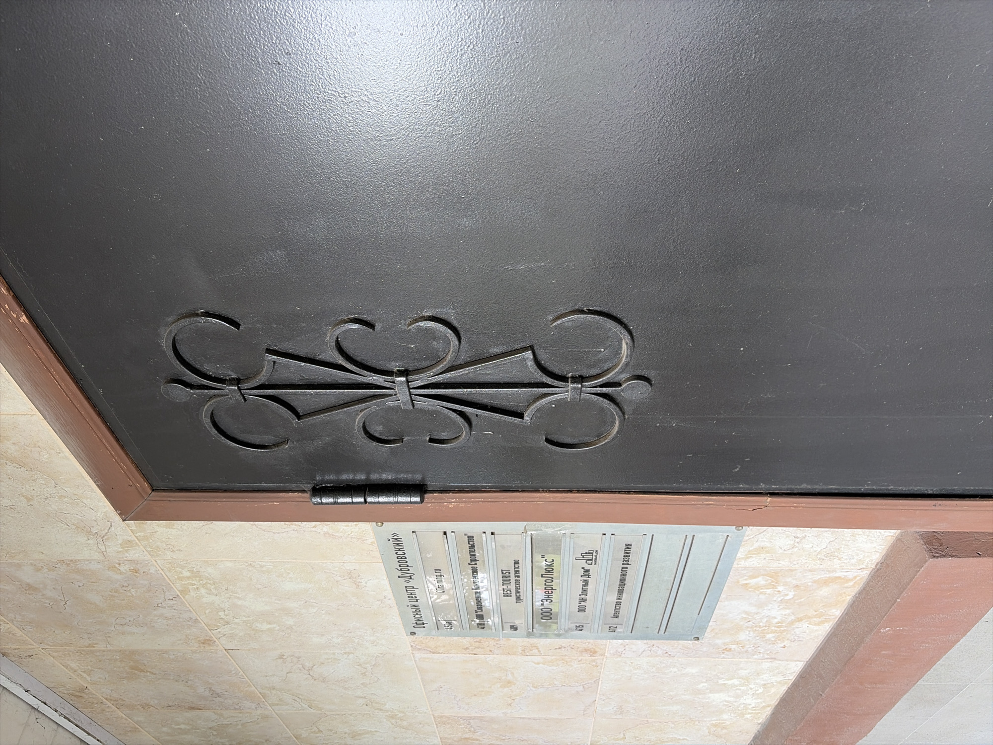 A close-up, eye-level shot shows a dark gray metal door with ornate black metal scrollwork in the center. To the left of the door is a beige tiled wall. Mounted on the wall to the left of the door is a metal directory listing various offices. The directory has a light-colored background and darker text. Some of the listed offices are "Офисный центр «Дубровский»", "QTraining.ru", "ООО "ЭнергоЛюкс"", and "Агентство инновационного развития". The directory is affixed to the wall with screws, one of which is visible. The door is hinged on the left side. The lighting suggests an indoor, possibly hallway, environment.