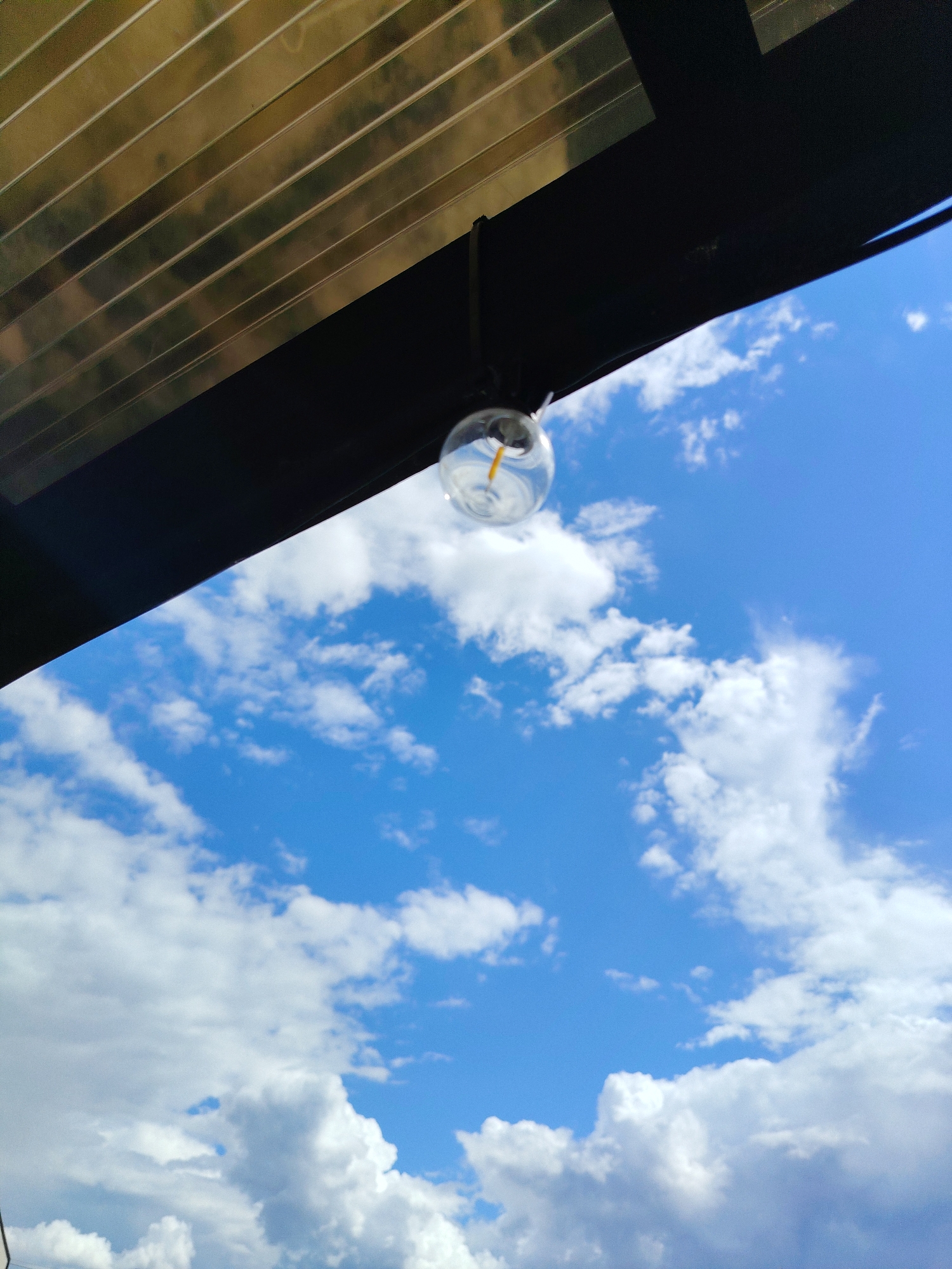 The image shows a view looking upwards towards a bright blue sky filled with fluffy white clouds. In the foreground, a dark overhang partially obscures the sky, with horizontal lines suggesting a textured ceiling or roof structure above it. Hanging from the overhang by a wire is a clear glass lightbulb. The lightbulb appears to be a decorative Edison-style bulb, with a visible filament inside. The sun is shining brightly, casting light on the clouds and the inside of the lightbulb. The scene conveys a sense of a bright, clear day, possibly outdoors on a patio or under a pergola. There is no visible text in the image.