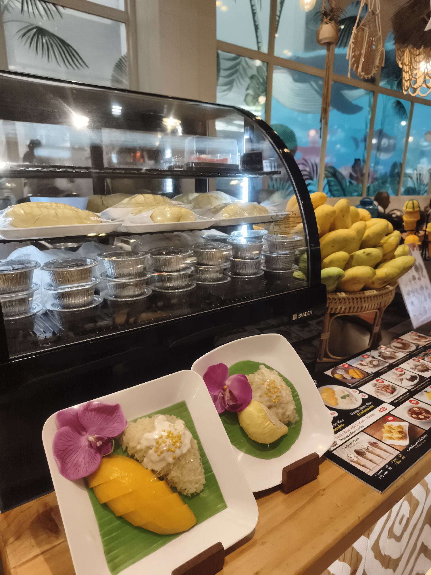A close-up, eye-level shot shows a refrigerated display case filled with food items, including individual portions of what appear to be desserts in foil containers and large, whole durians. In the foreground, two white plates each display a portion of mango sticky rice, garnished with a purple orchid and a slice of durian on one plate. Behind the display case, a basket overflows with ripe mangoes. To the right, a menu board showcases various food items and prices, including "Mango Sticky Rice with Durian." The background features a brightly lit, tropical-themed interior with large windows revealing a blue wall mural depicting aquatic life and palm trees. The overall setting suggests a food establishment, possibly a cafe or dessert shop, in a tropical location like Ko Samui District, Thailand.
