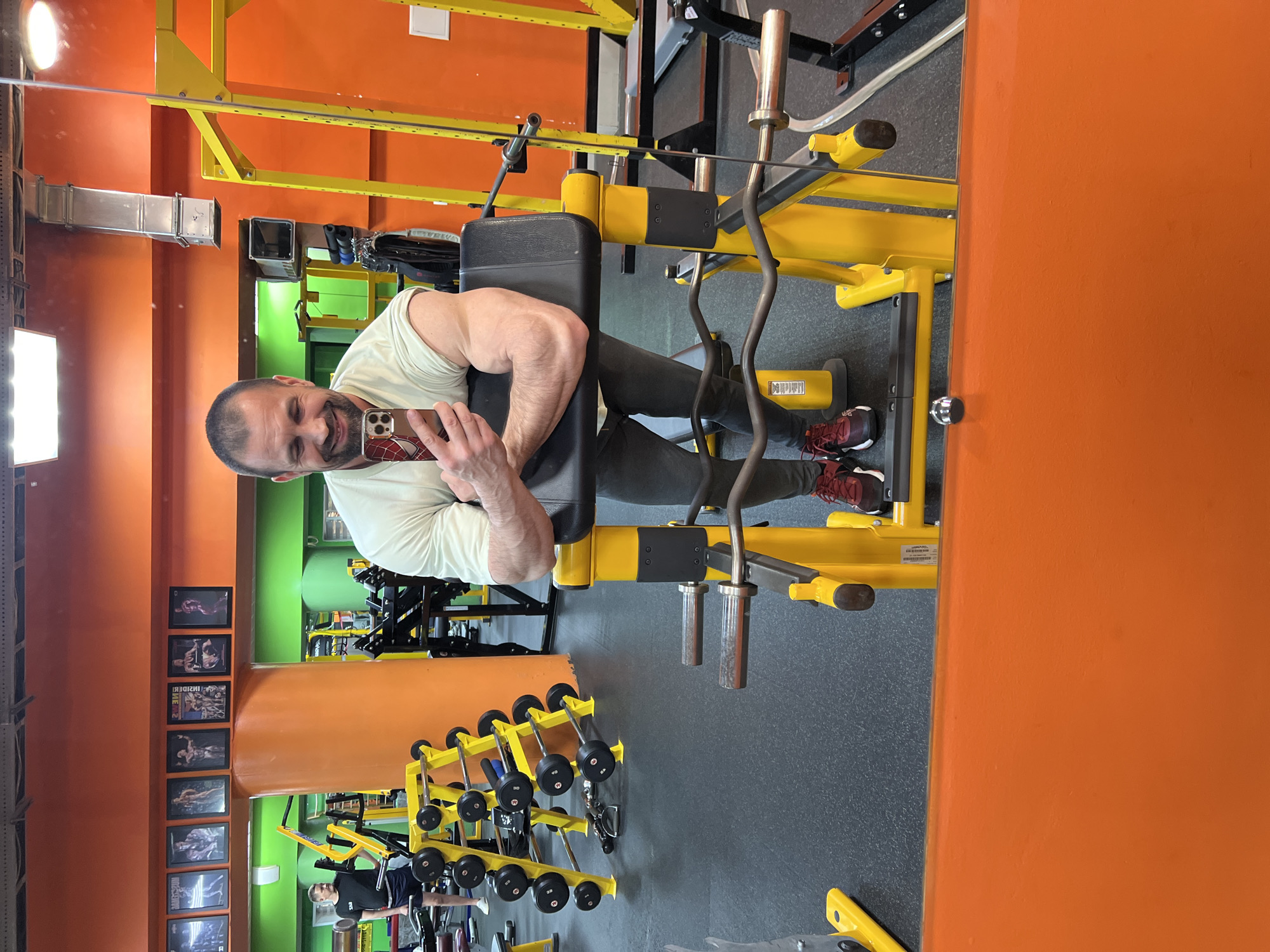 Here is a detailed description of the image:
**General Description:**
The image depicts a man taking a selfie in a gym using his smartphone. The gym appears to be well-equipped, with various weightlifting machines and equipment.
**People:**
* **Man:** An adult male is the primary subject of the image. He is holding a smartphone and taking a selfie. He is wearing a light-colored t-shirt and grey pants, with maroon sneakers. He has a bald head and is smiling, appearing to be in good physical condition.
**Objects:**
* **Gym Equipment:** A variety of weightlifting machines and equipment are visible, including a prone leg curl machine, barbells, and other machines. The equipment is predominantly yellow.
* **Smartphone:** The man is holding a smartphone, using it to take the selfie. The phone case seems to feature a Spider-man theme.
* **Mirror:** The image is a reflection in a mirror, as evidenced by the framing and perspective.
* **Wall décor:** Framed images adorn the orange wall.
**Actions and Interactions:**
* The man is taking a selfie in the mirror.
**Environment:**
* **Gym:** The setting is a gym.
* **Walls:** The walls of the gym are orange and green.
* **Lighting:** Artificial lighting is present, as indicated by the lamps.
**Location:**
The image was most likely taken in a gym located in Yoshkar-Ola, Russia.
I have included all the details I could perceive.