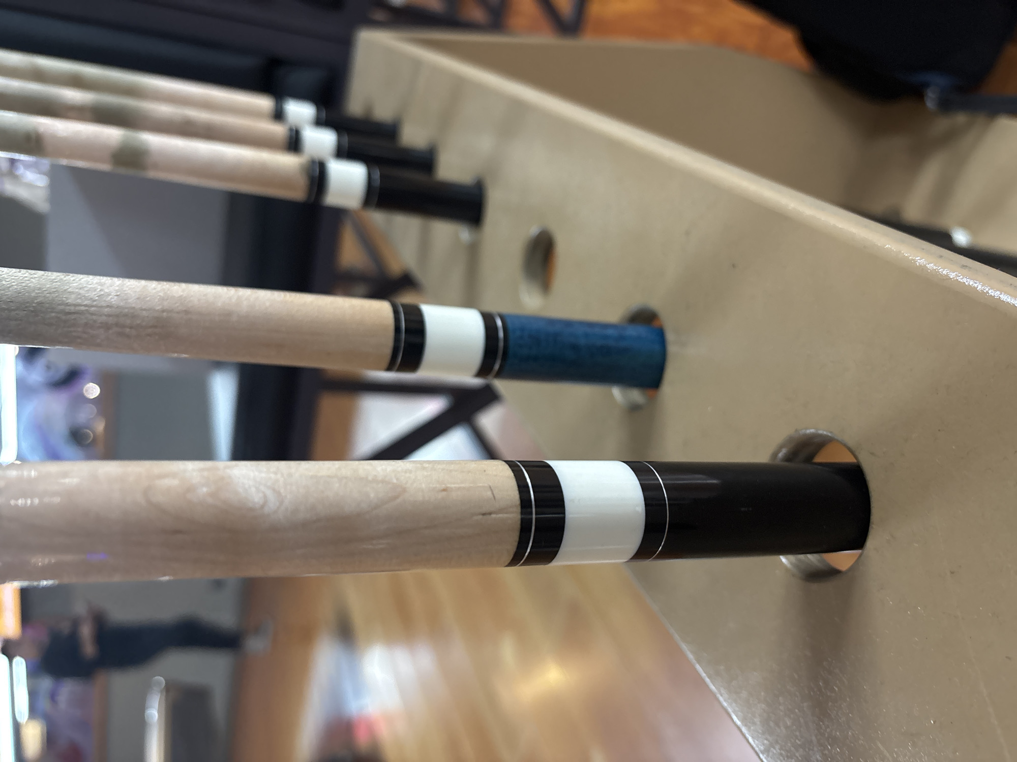 The image shows a close-up of several pool cues standing upright in a tan rack. The pool cues are made of wood and have distinct white and black bands, with one cue featuring a blue section. In the background, out of focus, a person wearing a dark shirt and pants is standing, possibly in a dimly lit establishment with wooden flooring and glowing screens visible. The scene appears to be indoors, likely a billiard hall or sports bar. The city of Kuta Selatan, Indonesia is not depicted in this image.