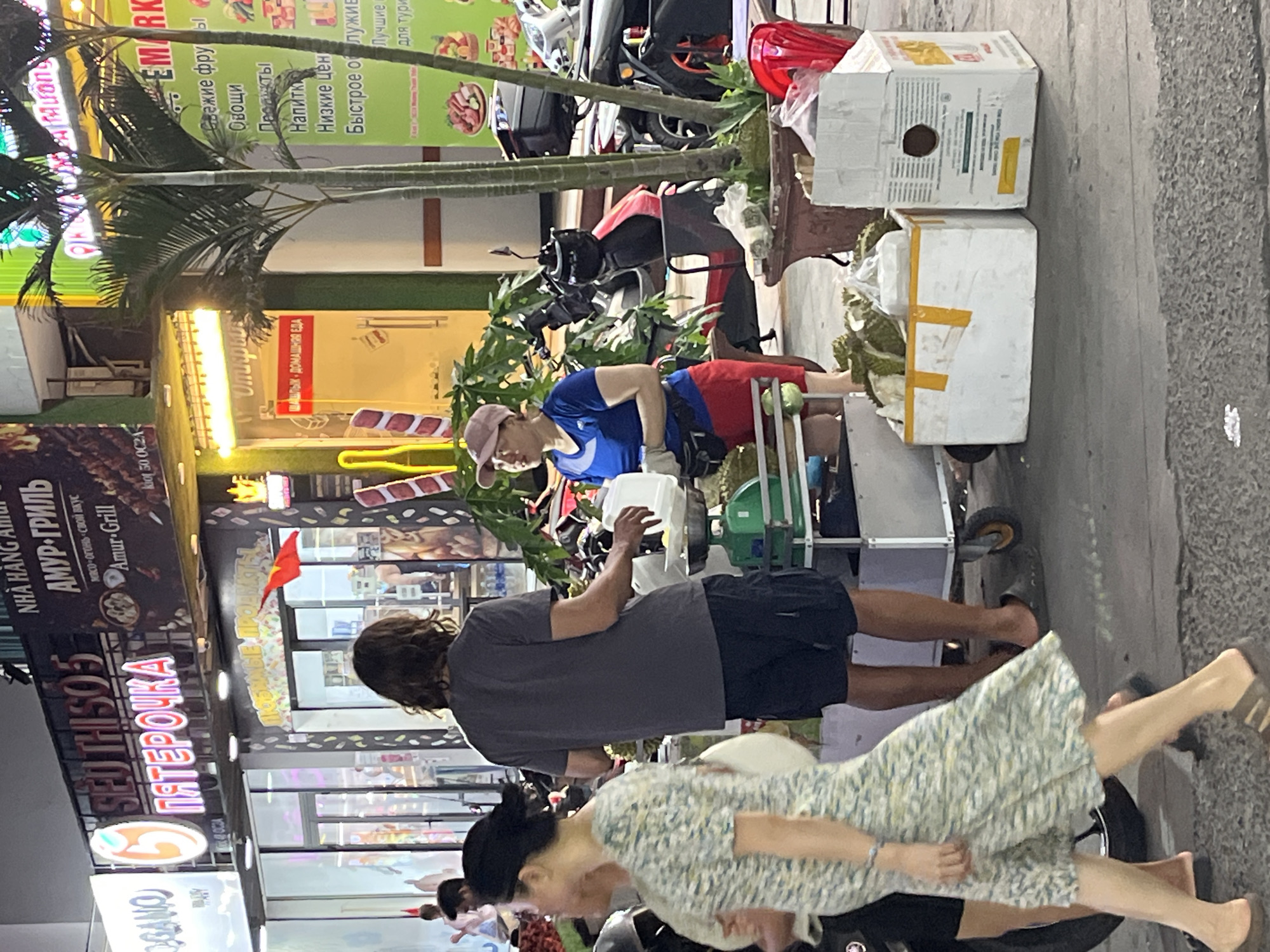 The image shows a street scene in Bac Nha Trang Ward, Vietnam. People are standing near a durian cart. One man is wearing a grey t-shirt and black shorts and is facing the cart. A woman in a floral dress is walking by. An Asian man sits by the cart and handles durians. Two white boxes are stacked up on the cart. Behind the cart, there are scooters. There is a palm tree on the left side of the image. The building on the left says "SEOTHI SO5" in red letters. There is an awning with the words "EMARK" in green and white. Vietnamese flags are hung on some of the buildings.