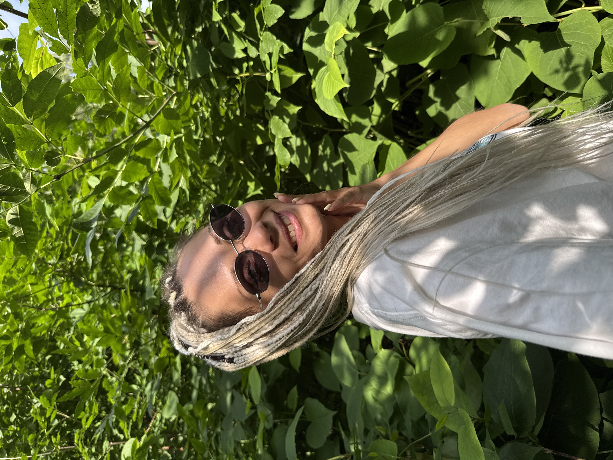A young woman with blonde braids and sunglasses is standing outdoors in front of a dense green foliage background. She is smiling and resting her hand on her cheek. The sun is shining through the leaves, casting shadows on her face and white t-shirt. The location appears to be a natural setting, possibly a park or wooded area, and is not identifiable as being in Rusanovo, Russia, or any specific city.