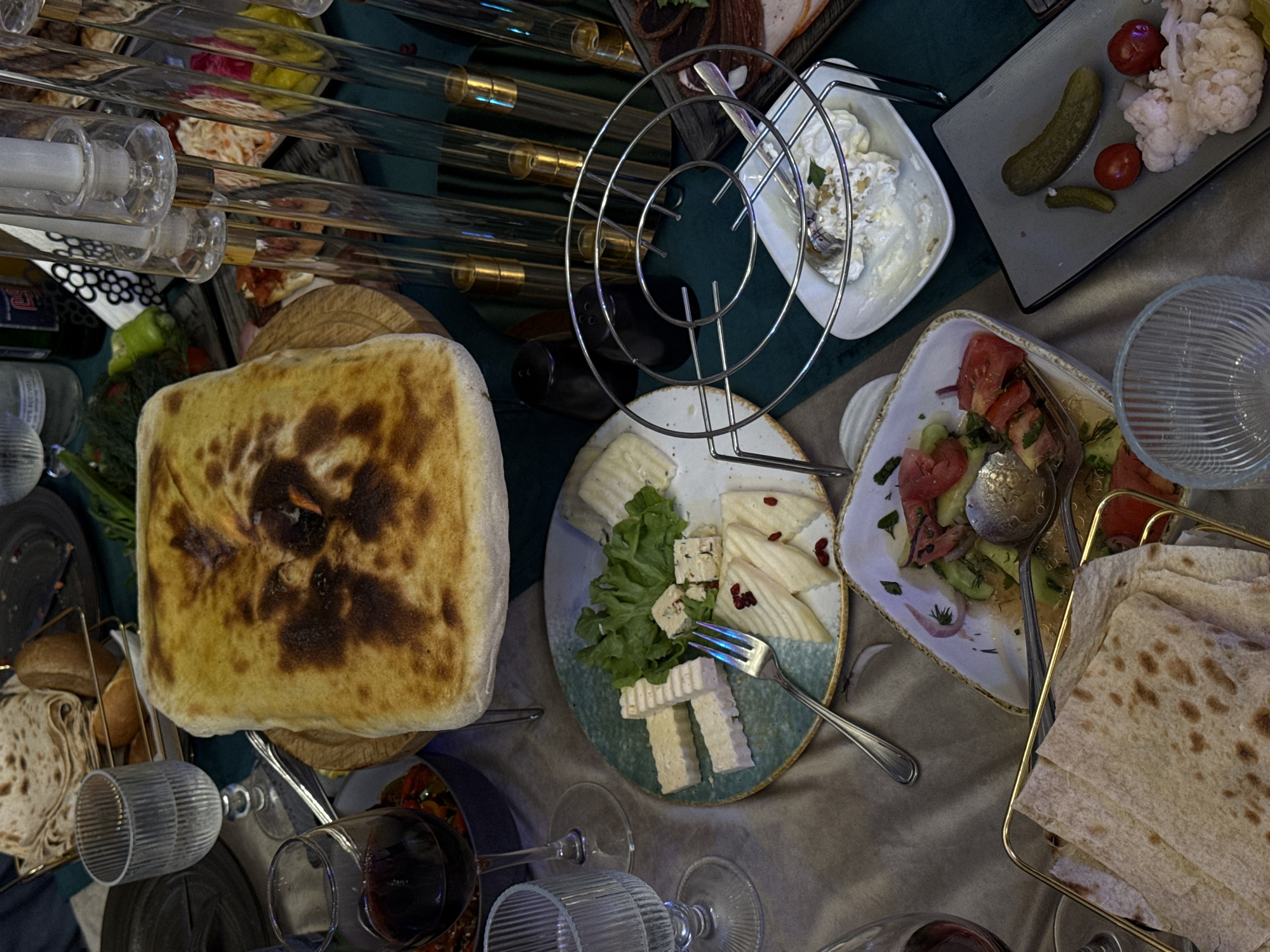 The scene is a table laden with various dishes, likely photographed in Yerevan, Armenia, given the presence of traditional Armenian cuisine.
Objects:
* A large, golden-brown flatbread (likely a type of lavash or matnakash) dominates the left side of the frame.
* Assortment of cheeses on a decorative plate.
* A plate with sliced cucumbers, tomatoes, and herbs (likely a salad).
* A dish containing white creamy food with nuts.
* Pickled vegetables (cucumbers, tomatoes, cauliflower).
* Stacks of thinner flatbread (another type of lavash).
* Wine glasses, some filled with red wine.
* Metal serving stands are visible.
* Candles in glass holders.
The setting suggests a celebratory or gathering atmosphere, possibly in a restaurant or private dining room.