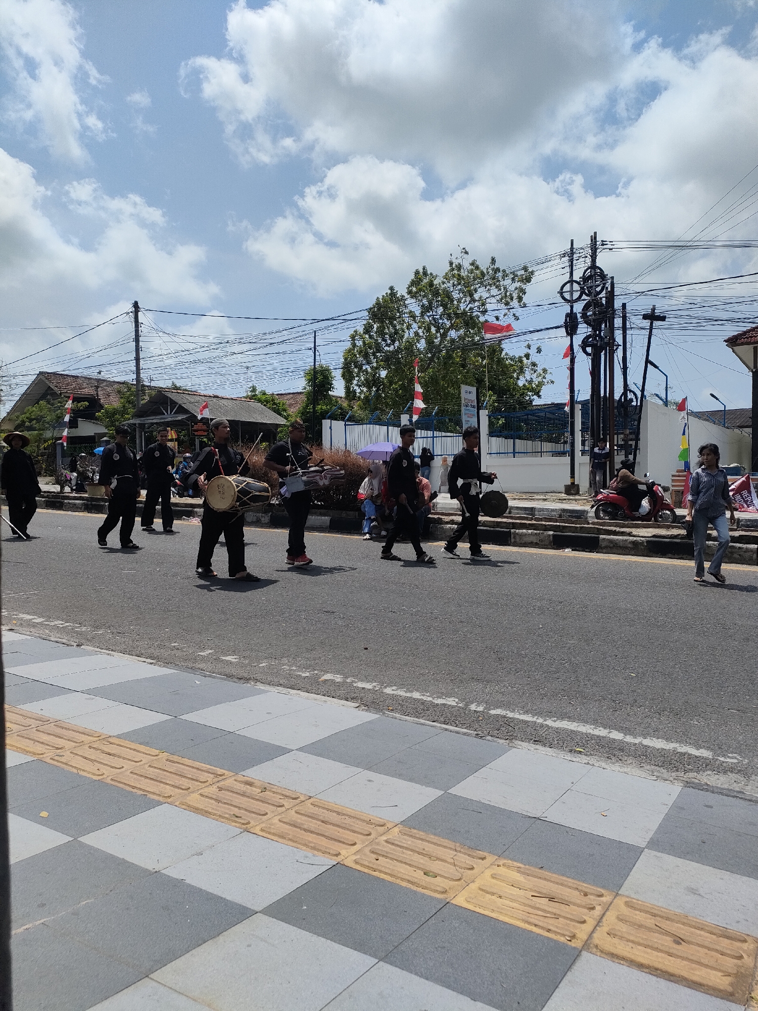 In Sungailiat, Indonesia, a parade is taking place on a street. A group of individuals in black uniforms are walking in procession, some carrying musical instruments like drums and what appears to be a violin. They are accompanied by spectators on the side of the road. Red and white Indonesian flags are visible, suggesting a national celebration. The street is paved with asphalt, and a sidewalk with tactile paving for the visually impaired is present. Buildings and trees line the street, and utility poles with numerous wires are prominent features of the urban environment. The sky is partly cloudy.
