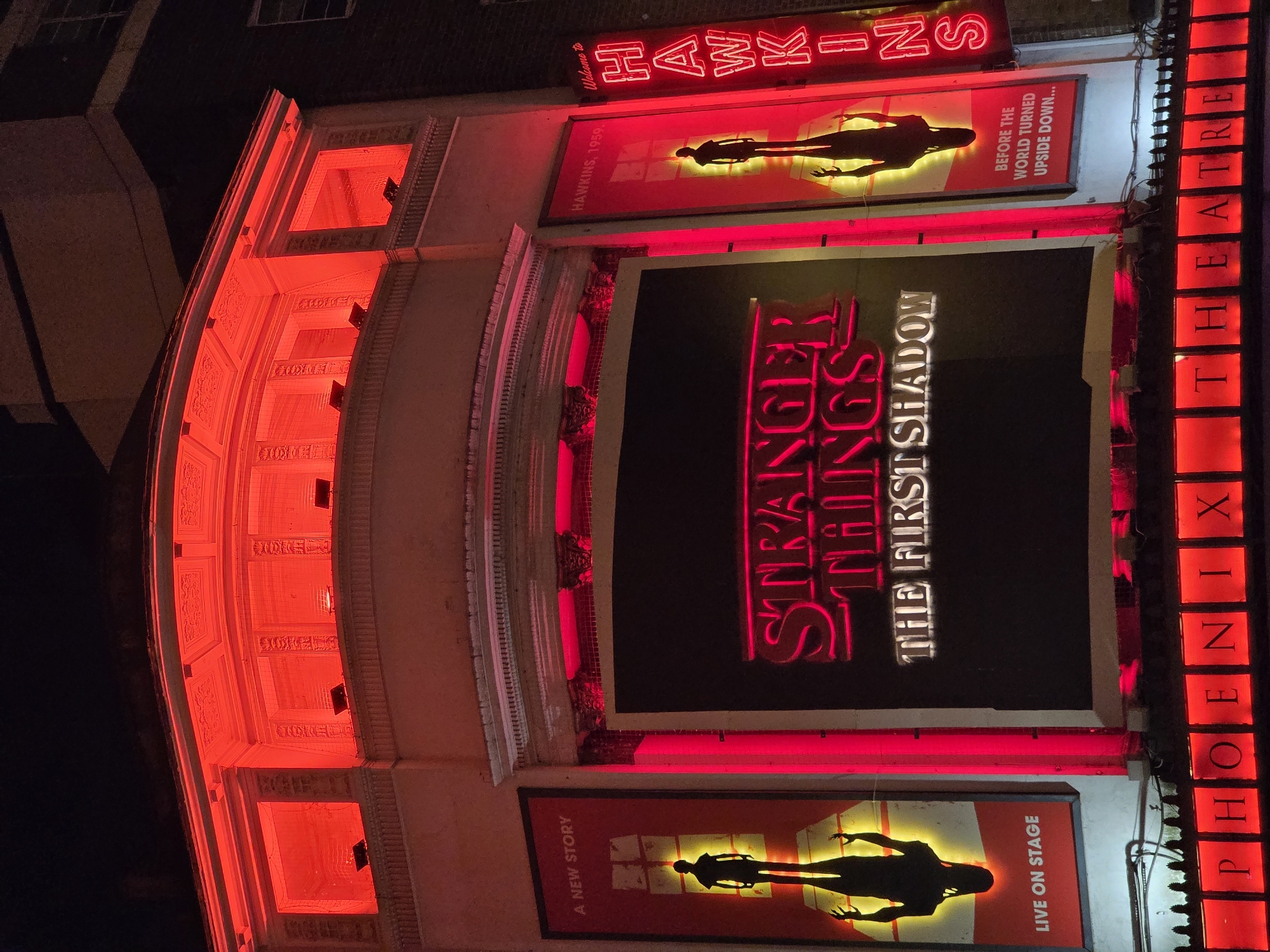The image captures the exterior of the Phoenix Theatre in London, United Kingdom, bathed in red lighting. Large signs promote the play "Stranger Things: The First Shadow." There are two smaller posters of what appears to be a figure standing near a bright glowing place. The name "HAWKINS" is illuminated in neon above one of the posters, with the phrase "Welcome to HAWKINS." A vertical sign displaying "PHOENIX THEATRE" is on the right side of the photograph. The theater's facade is visible with details like arched windows and intricate architectural elements, illuminated in red. The setting appears to be at night.