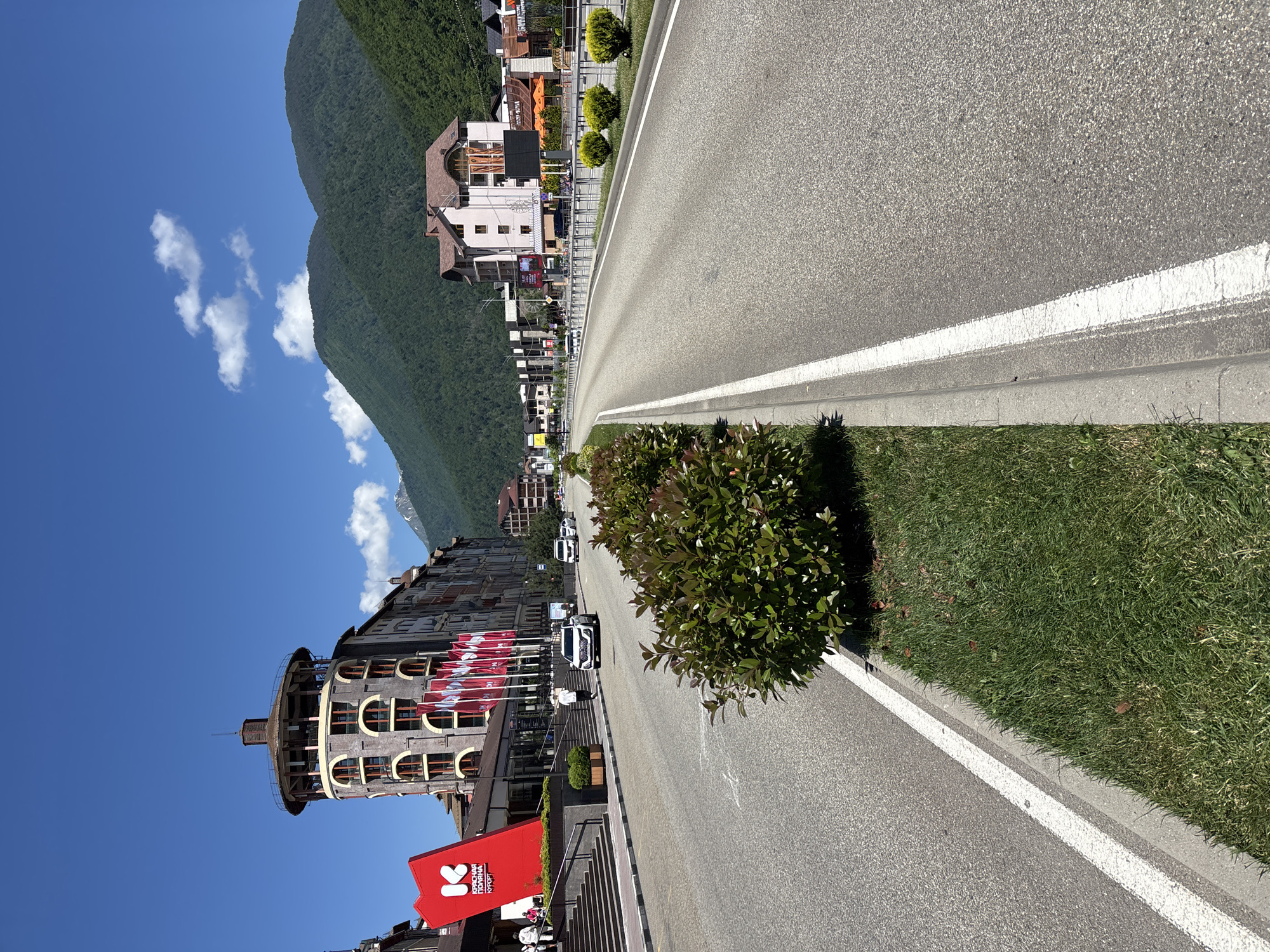 The image depicts a sunny day in Estosadok, Russia, with a clear blue sky and scattered clouds. A paved road runs through a town nestled at the base of a lush, tree-covered mountain. The road is lined with green grass and manicured bushes.
On the left side of the road, a multi-story building with a curved, tower-like section is prominently featured. This building has numerous arched windows and is adorned with red flags displaying white text. Nearby, a large red banner with white text is visible, seemingly advertising a local event or establishment. Other buildings, typical of a resort town, are visible further down the road, with hotels and shops lining the street. Several cars are driving on the road, including white SUVs and a white car with a dark roof. A few people are visible on the sidewalks and near the buildings, dressed in casual clothing.
The scene suggests a vibrant tourist or recreational area. The clear weather and bright sunlight indicate it is daytime, likely late morning or early afternoon. The mountains in the background and the architecture of the buildings suggest a location in a mountainous region known for outdoor activities. The overall mood conveyed by the image is bright and cheerful, with a sense of bustling activity in a scenic environment.
