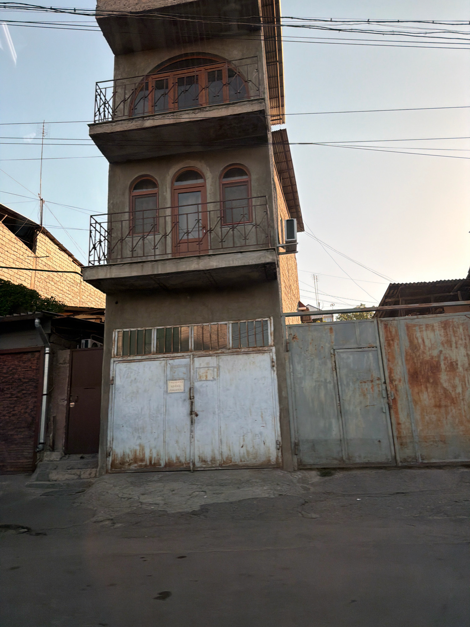 The image depicts a tall, narrow building with three stories, located in Yerevan, Armenia. The building has a concrete facade and features arched windows on the second and third floors, with wrought-iron balconies. The ground floor has a large set of metal garage doors. To the left of the garage doors is a brown, single door. To the right of the garage doors is a large, rusty metal gate. The ground in front of the building is a rough, asphalted surface. Overhead, numerous electrical wires are visible against a pale sky. The overall scene suggests a residential or mixed-use area within the city.