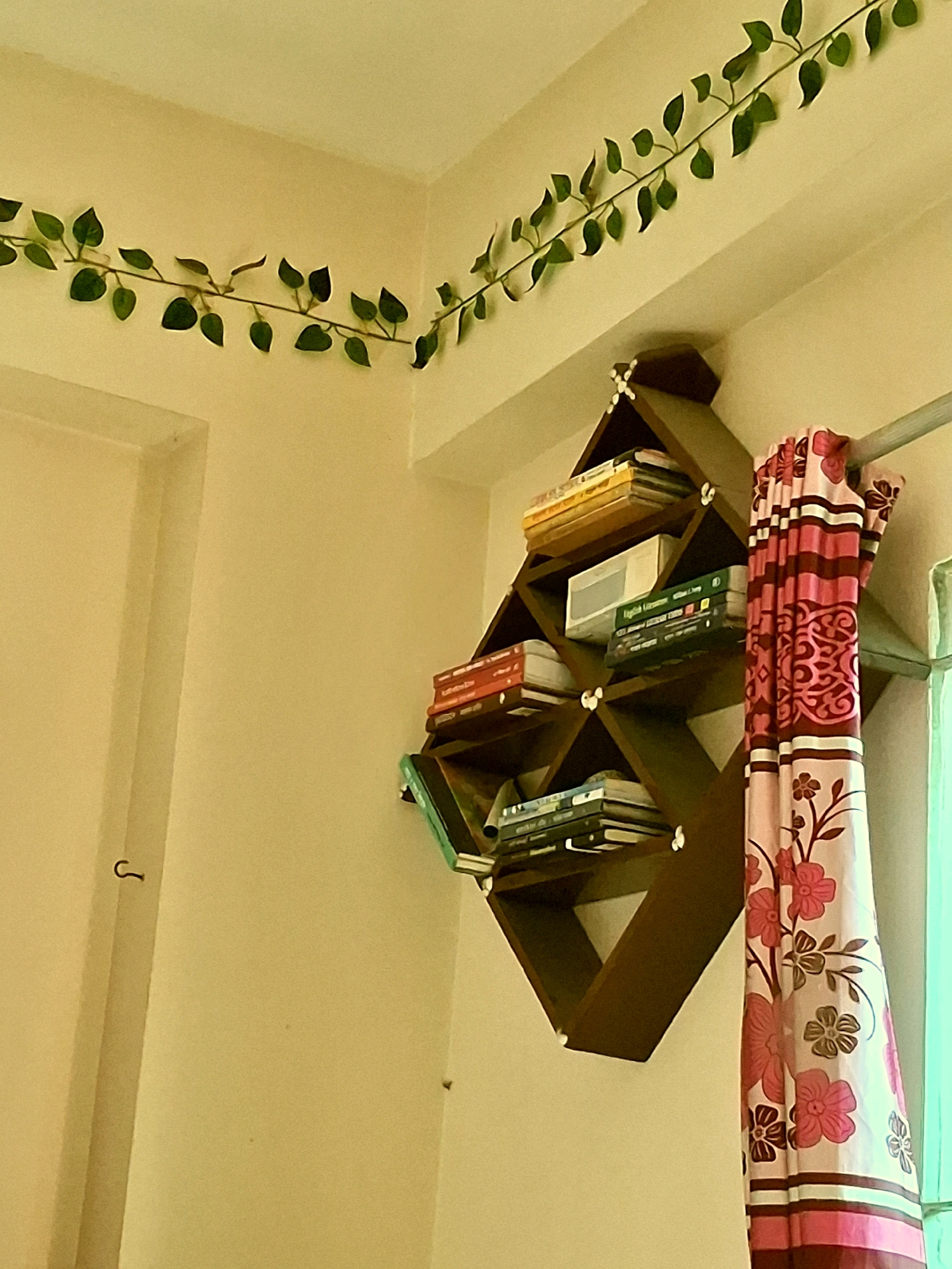 The image shows an interior scene with a wall-mounted wooden bookshelf filled with books. The bookshelf has a unique geometric design with diamond-shaped compartments. A pink and white floral curtain hangs to the right of the bookshelf, partially obscuring a window. Green artificial vine decor is draped along the top corner of the wall. The walls are painted a light cream color. A hook is visible on the left side of the wall. The scene appears to be a residential interior in Dhaka, Bangladesh.
