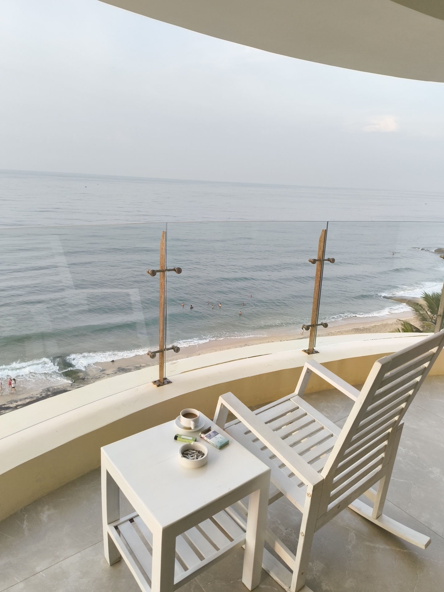 An exterior shot captures a balcony in Phu Thuy Ward, Vietnam, overlooking a serene beach and the ocean. The balcony is furnished with a white slatted rocking chair and a small white side table. On the table, a white teacup with a saucer holds a dark liquid, likely coffee or tea. Beside it rests a pack of cigarettes and a green lighter, with an ashtray filled with cigarette butts. The glass railing of the balcony is partially visible, with wooden posts supporting it. In the background, the calm ocean stretches to the horizon under a hazy sky. The beach is sandy, with gentle waves lapping at the shore. A few people are visible in the water, swimming or wading. Lush green palm trees are dotted along the coastline. The overall atmosphere is peaceful and relaxed, suggesting a quiet morning or late afternoon by the sea.