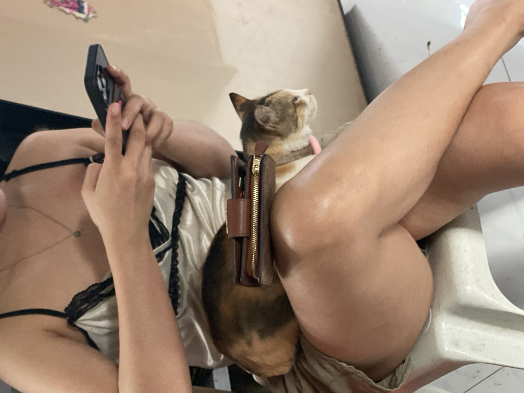 An indoor scene features a person relaxing with a cat and a smartphone. The person, whose upper body and legs are visible, is wearing a light-colored top with black lace detailing, possibly sleepwear or lingerie. They are holding a black smartphone in both hands, seemingly engrossed in it. Their legs are crossed and bare, suggesting a casual and comfortable setting.
A calico or tortoiseshell cat with brown, black, and white fur is nestled between the person's legs, wearing a pink collar. The cat's head is tilted upwards, looking towards an unseen point above, possibly at something interesting or just in a contemplative pose. A brown leather wallet with a gold zipper is placed upright between the cat's back and the person's left leg.
The background is a simple, light-colored wall, possibly part of a home interior. A white plastic chair or stool is partially visible supporting the person's lower body. The overall atmosphere is one of domestic comfort and relaxation. The image lacks specific time-of-day indicators but suggests an intimate, personal moment. There is no visible text. This scene takes place in Davao City, Philippines.