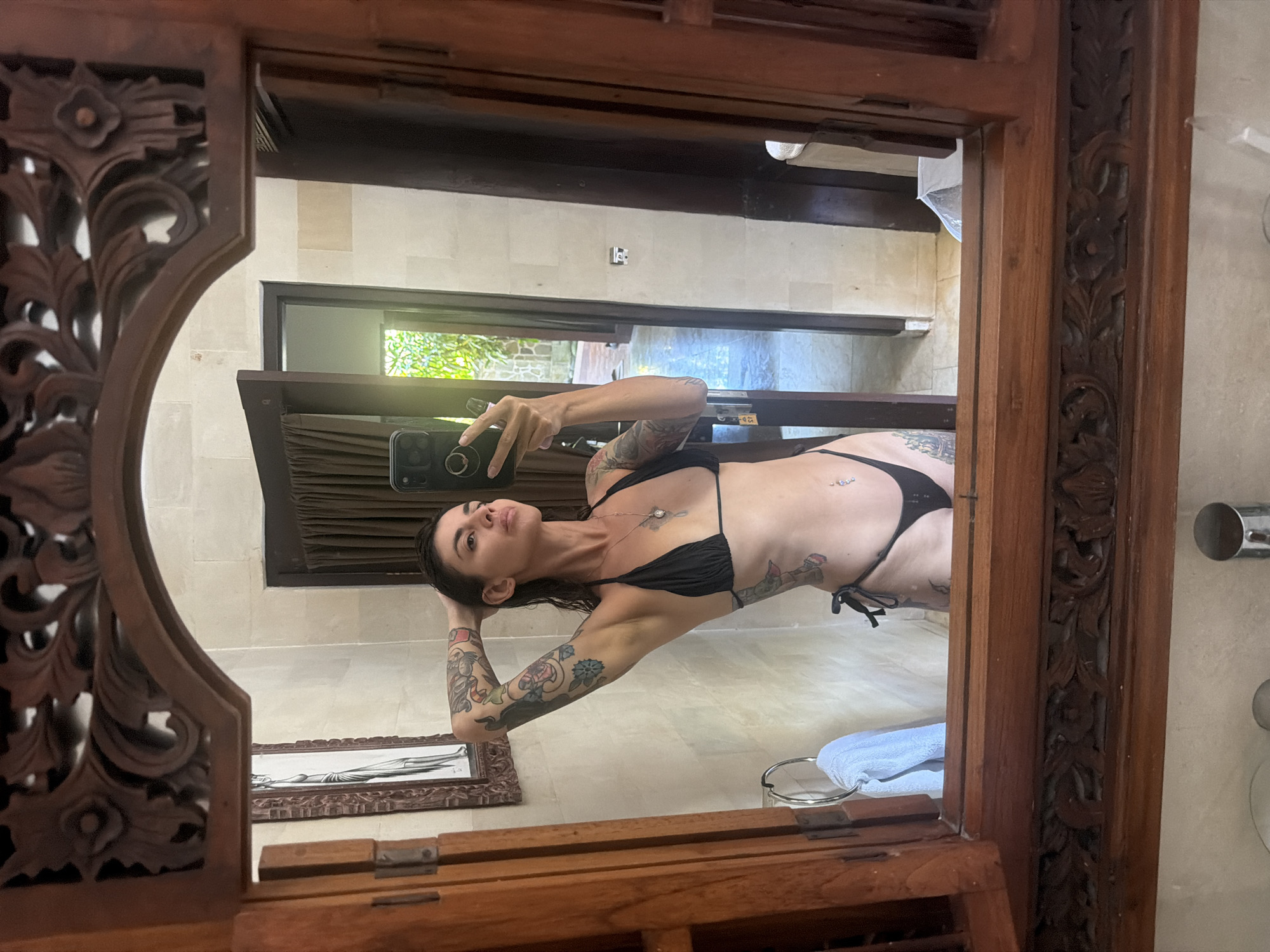 A woman with extensive tattoos on her arms and torso, wearing a black bikini, is taking a mirror selfie in a bathroom. She has her right hand behind her head, and her left hand is holding a smartphone. The bathroom features a large wooden framed mirror, tiled walls, and a door leading to another area. A towel hangs on a ring to the left of the mirror. The scene suggests a warm, tropical environment, consistent with a location like Blahbatuh, Indonesia.