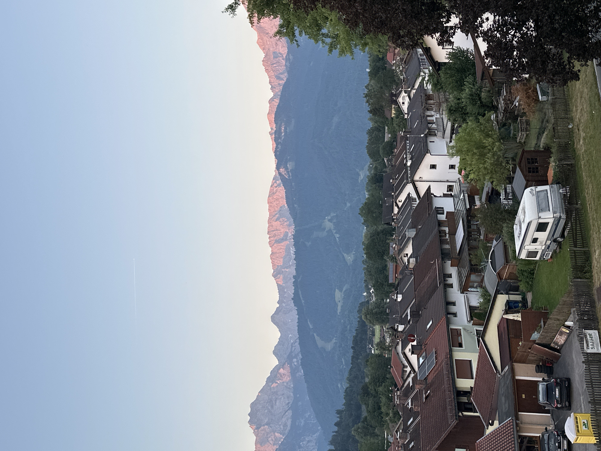 The image shows a residential area in Garmisch-Partenkirchen, Germany, with a mountain range in the background. The sky is clear and blue with a faint contrail from an airplane. The sun is low, casting a warm, pinkish glow on the mountain peaks. In the foreground, there are rows of houses with red-tiled roofs. Some houses have solar panels on their roofs. A few cars are parked in driveways. There is a caravan parked on a grassy area next to a small shed. A yellow portable toilet is also visible. Trees and vegetation surround the houses. The overall scene depicts a quiet, suburban setting with a dramatic natural landscape.