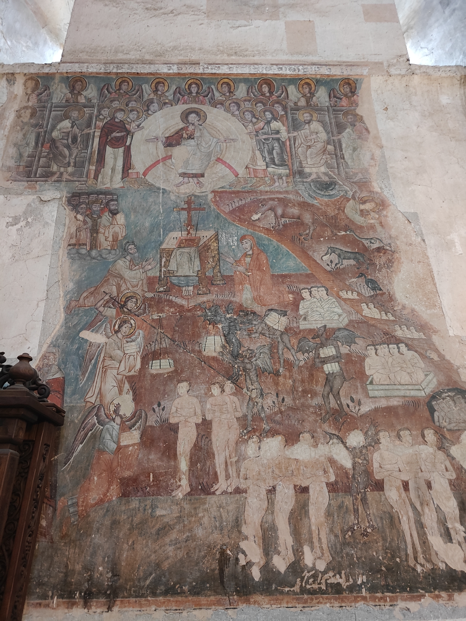 This image shows a fresco painting on a wall. The painting is weathered, with some sections faded or damaged.
The top portion depicts a central figure surrounded by an aureole, possibly Christ, with attendant figures including angels behind and several people below.
The middle section shows several scenes including:
1. A cross on top of what appears to be a table.
2. An angel holding a scale.
3. What looks like the depiction of both Heaven and Hell.
The bottom portion of the mural depicts multiple figures standing and walking.
Given the location (Pkhundavi, Georgia), this is likely a religious scene from a Georgian Orthodox Church. The subject may be The Last Judgement.