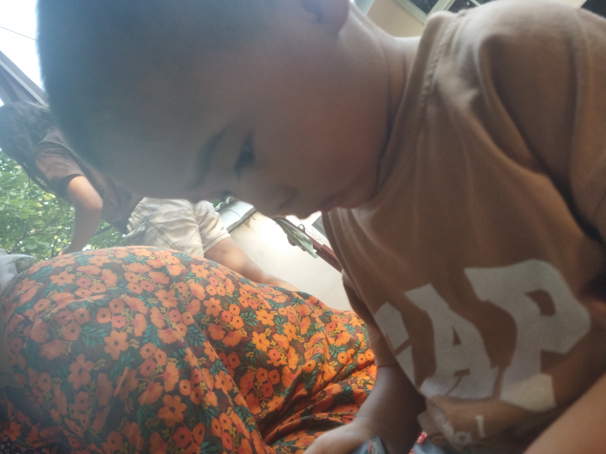 In this close-up, candid shot, two individuals are partially visible, suggesting an informal gathering. The foreground is dominated by a child, whose head and shoulder are prominently featured, looking downwards. The child is wearing a tan t-shirt with "GAP" written in large white letters across the chest. To the left of the child, a portion of another person is visible, wearing a light-colored shirt. Further left, a third person is visible, with dark hair and a tan shirt, partially obscured by the child and the overall framing of the image. The background shows a hint of greenery, suggesting an outdoor setting, possibly during the daytime given the bright lighting. The arrangement of people and the casual attire imply a relaxed, everyday moment. The focus is tightly on the child and their t-shirt, with the other individuals providing context to the scene. The overall impression is one of intimacy and personal connection, captured in a fleeting moment.