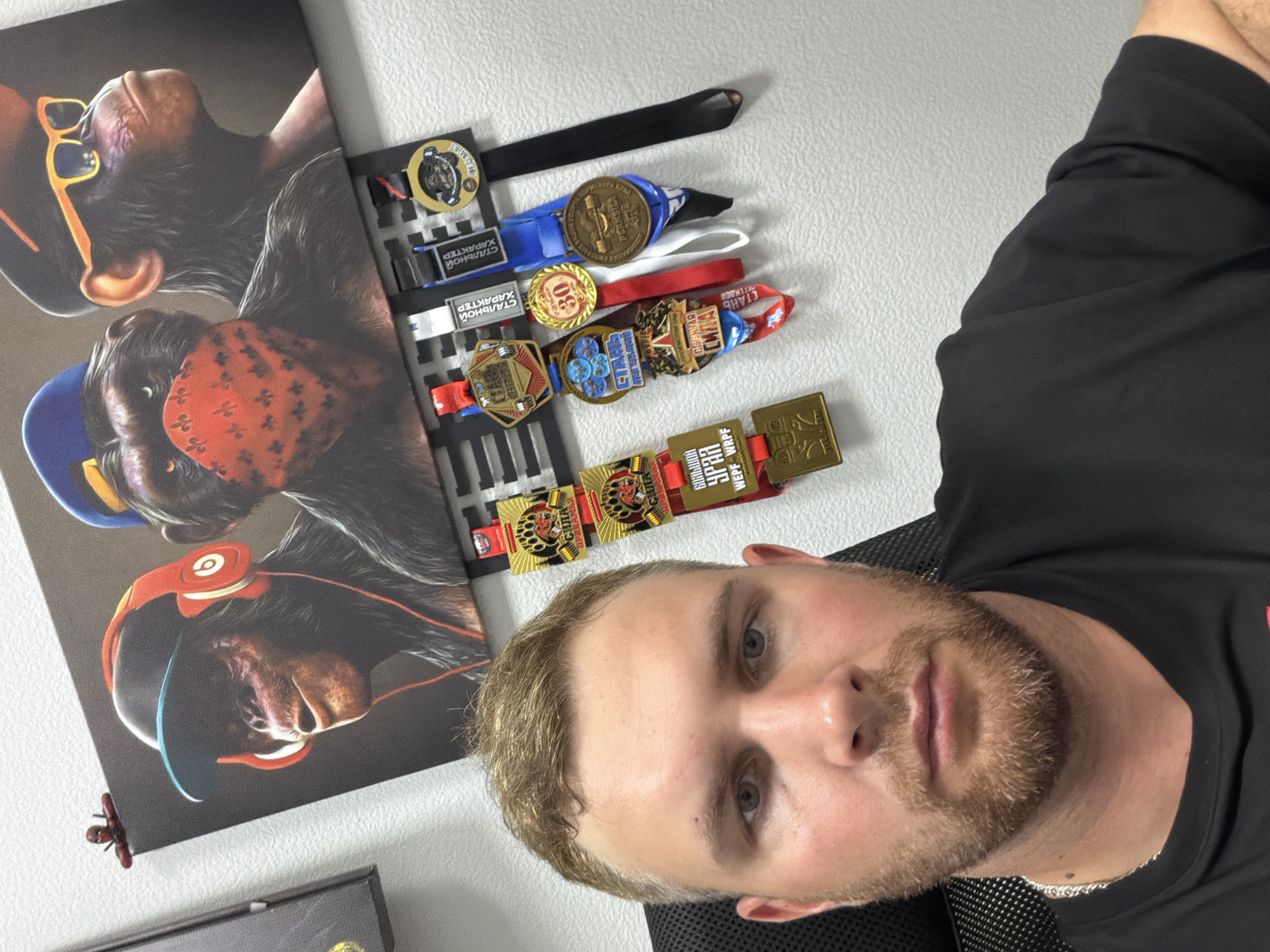 The image depicts a man with short blonde hair and a beard, wearing a black t-shirt and a silver chain, seated in a black office chair. He is looking directly at the camera, with his face and chest filling the lower half of the frame. Behind him, on a white wall, hangs a picture of three chimpanzees wearing accessories like headphones, a bandana, and sunglasses. Below the picture, a medal display rack holds several medals with colorful ribbons. One of the medals features an image of a bear and text that appears to be in Russian, reading "СИЛА ПРИКАМЬЯ". Another medal has the number "30" on it. The overall environment suggests an indoor setting, possibly an office or a personal space. There is no information within the image that specifies a location within the city of Chaikovsky, Russia.