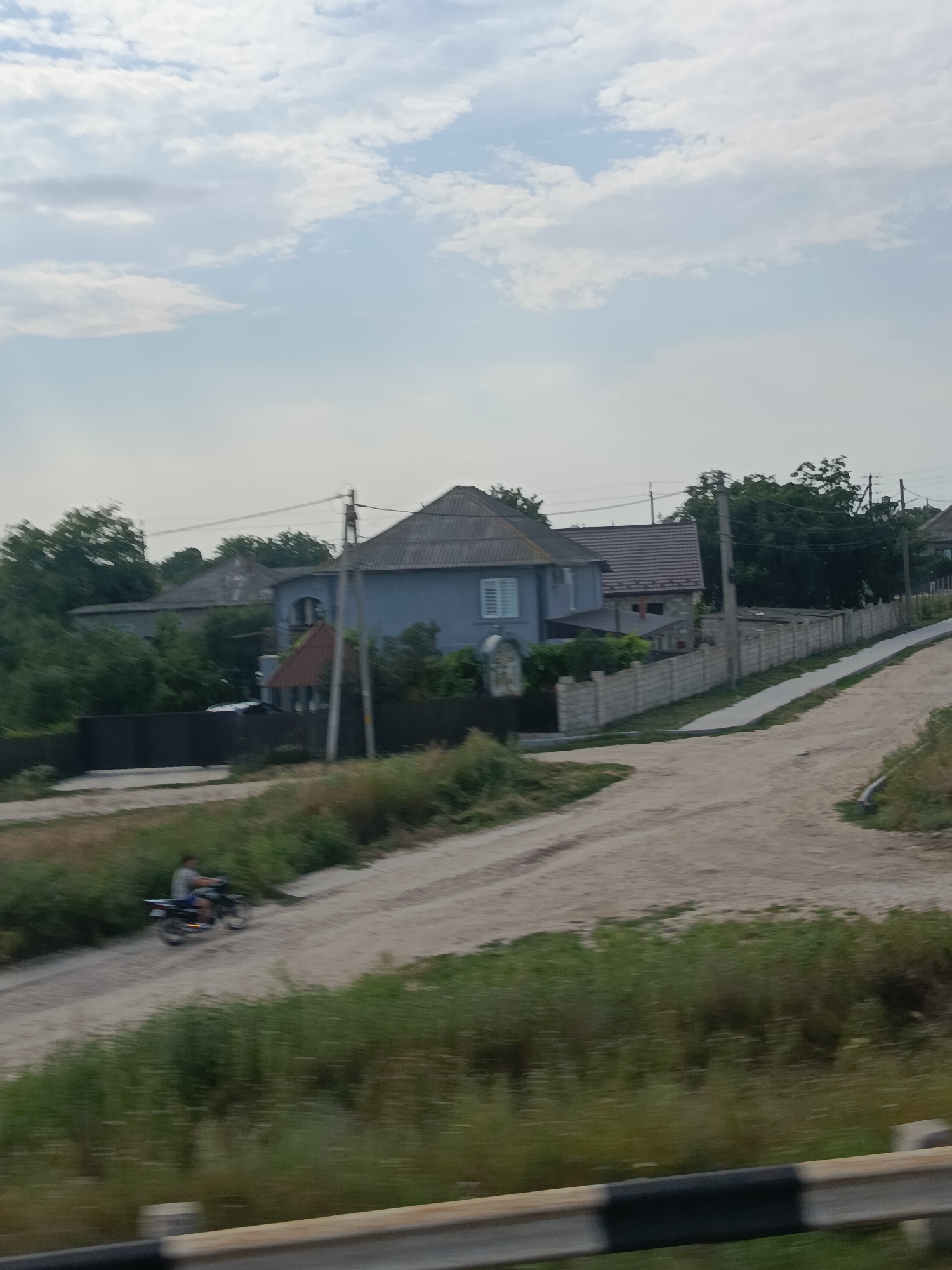 This image captures a rural scene in Razeni, Moldova. A dirt road winds through the landscape, bordered by lush green grass and trees. In the background, several houses with grey-tiled roofs are visible, nestled amongst the foliage. A concrete fence separates some of the properties from the road.
The activity depicted is a person riding a motorcycle on the dirt road. The rider is wearing a grey shirt and shorts, and the motorcycle is dark in color. The scene suggests a casual daytime activity, likely in the afternoon given the lighting. The sky is partly cloudy, with a mix of white clouds and blue sky, indicating fair weather.
Notable details include the distinct architectural style of the houses, characteristic of rural Eastern European settings. The presence of power lines suggests that these are inhabited properties. The overall impression is one of a peaceful, everyday moment in a countryside setting. There is no visible text in the image.