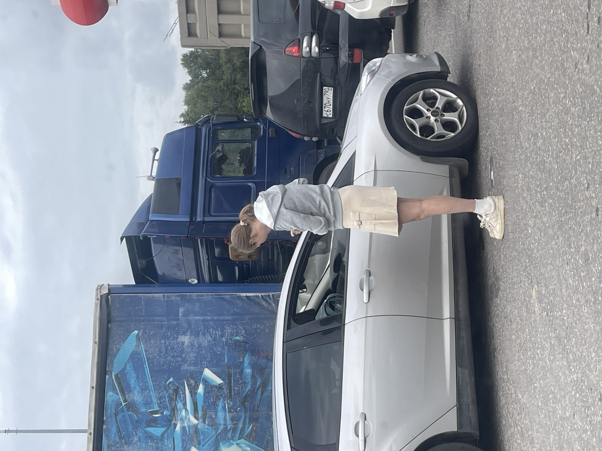 A young woman with blonde hair tied in a ponytail is leaning into the open driver's side window of a silver car. She is wearing a gray hooded sweatshirt and a light-colored skirt. She appears to be looking down at something inside the car. The car is parked on an asphalt surface. Behind the silver car are other vehicles, including a dark-colored SUV and a large blue truck with a trailer. The trailer has a large graphic of blue, angular shapes, possibly representing crystals or ice. The scene appears to be outdoors, with a cloudy sky visible in the background. The overall impression is of a casual moment captured on a possibly overcast day. There is text visible on the license plate of the dark SUV, which reads "C670HY 790". The location context provided is Istra, Russia.