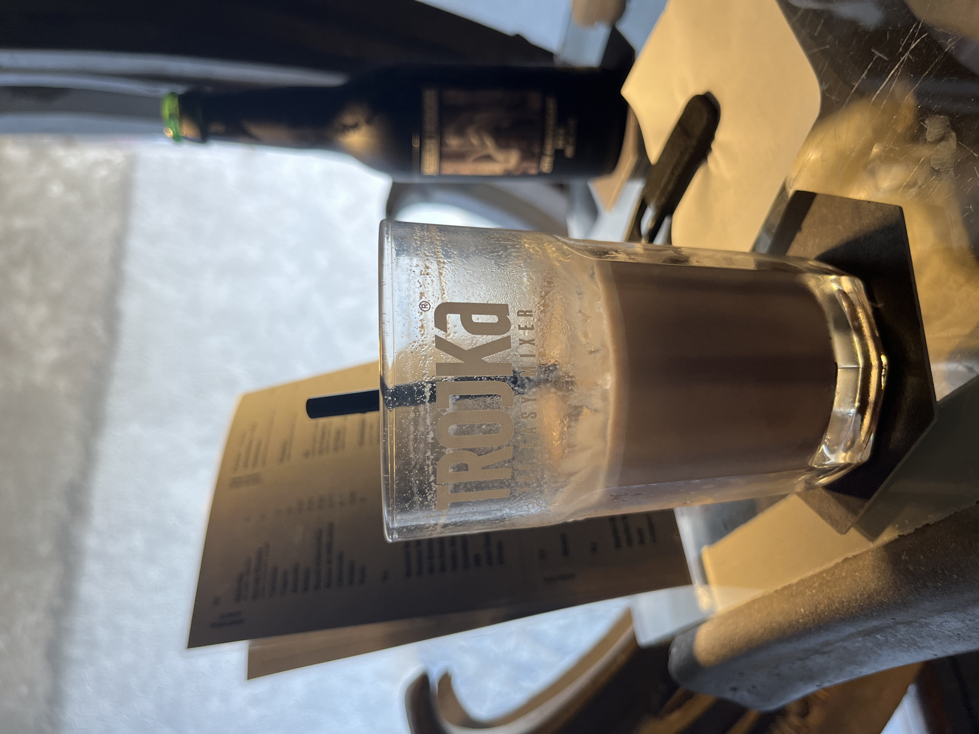 The image depicts a close-up, eye-level view of a glass containing a chocolate-colored beverage, likely hot chocolate or a similar drink. The glass has the word "TROJKA" emblazoned on it in white lettering, with "MIXER" below. A black straw is inserted into the drink. The glass is set on a black coaster, which rests on a glossy, reflective table.
In the background, slightly out of focus, is a brown menu or pamphlet propped open. To the right of the glass, a dark glass bottle, possibly containing a beverage, stands upright. The bottle has a label with an image on it. The bottle has a green cap. The overall environment appears to be indoors, with soft, diffused lighting, suggesting a cafe or restaurant setting. The background outside of the window is blurred and appears to be a bright, possibly overcast or snowy day. The location is identified as Epagny, Switzerland. There are no people visible in the image.