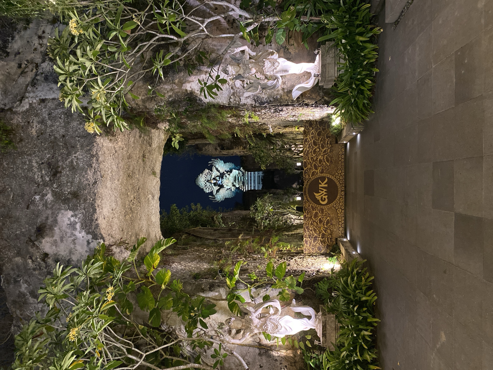 This image depicts an outdoor scene at night, likely in Kuta Selatan, Indonesia. The foreground features lush green foliage and flowering plants cascading over rough, rocky terrain. To the right, a paved pathway made of large, dark gray tiles is illuminated by small uplights. Adjacent to the pathway is a decorative panel with intricate, golden patterns on a dark background. The text "GWK" is visible within a circular emblem on this panel.
Further into the scene, the pathway leads towards a prominent statue of a figure, possibly mythological or religious, illuminated from below. This statue appears to be carved from white stone and is adorned with delicate details. Beyond this, in the background, a larger, more complex statue is visible, also illuminated, which appears to be a winged deity or mythical creature. The overall setting suggests a cultural or tourist attraction, possibly a temple complex or a monument, given the grand statues and decorative elements. The lighting creates a dramatic and somewhat mystical atmosphere. The presence of statues, intricate carvings, and tropical vegetation are strong indicators of the location's cultural heritage and geographical context. The image seems to capture a serene moment after dark, with artificial lighting highlighting the artistic and architectural features.