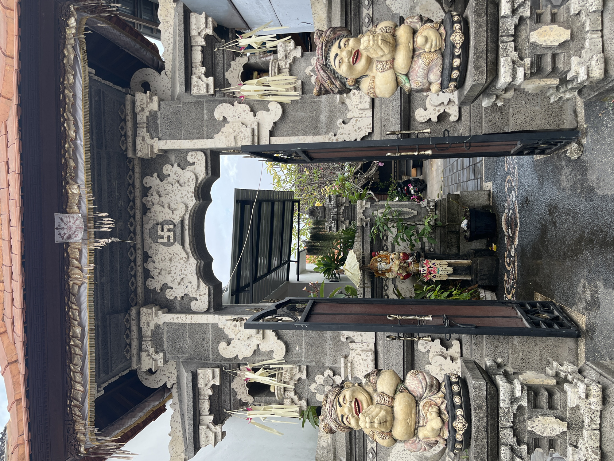 The image shows an ornate stone gateway in Kuta Selatan, Indonesia, likely the entrance to a temple or a traditional Balinese compound. The gateway is constructed of grey stone blocks and intricate carvings, topped with a terracotta tile roof. Two cheerful, plump statues flank the entrance, seated on stone pedestals. These figures, often seen in Balinese culture, appear to be made of weathered stone or concrete and wear traditional head coverings and patterned garments. They are depicted with wide smiles and clasped hands in a welcoming gesture.
Hanging from the gateway are decorative elements, including what appear to be palm leaf decorations and shimmering golden fabric trim. Above the archway, a Swastika symbol is prominently displayed, a common motif in Hindu iconography. Beyond the open metal gates, a pathway paved with patterned tiles leads further into the compound. Small shrines and potted plants are visible, contributing to the spiritual and natural atmosphere of the location. A statue of a deity, possibly Ganesha due to its elephant-like features, stands to the right, adorned with an umbrella-like canopy. A parked motorcycle is partially visible in the background, suggesting this is a lived-in space rather than a purely ceremonial one. The ground appears damp, indicating recent rain. The sky is overcast, casting a soft, diffused light on the scene.