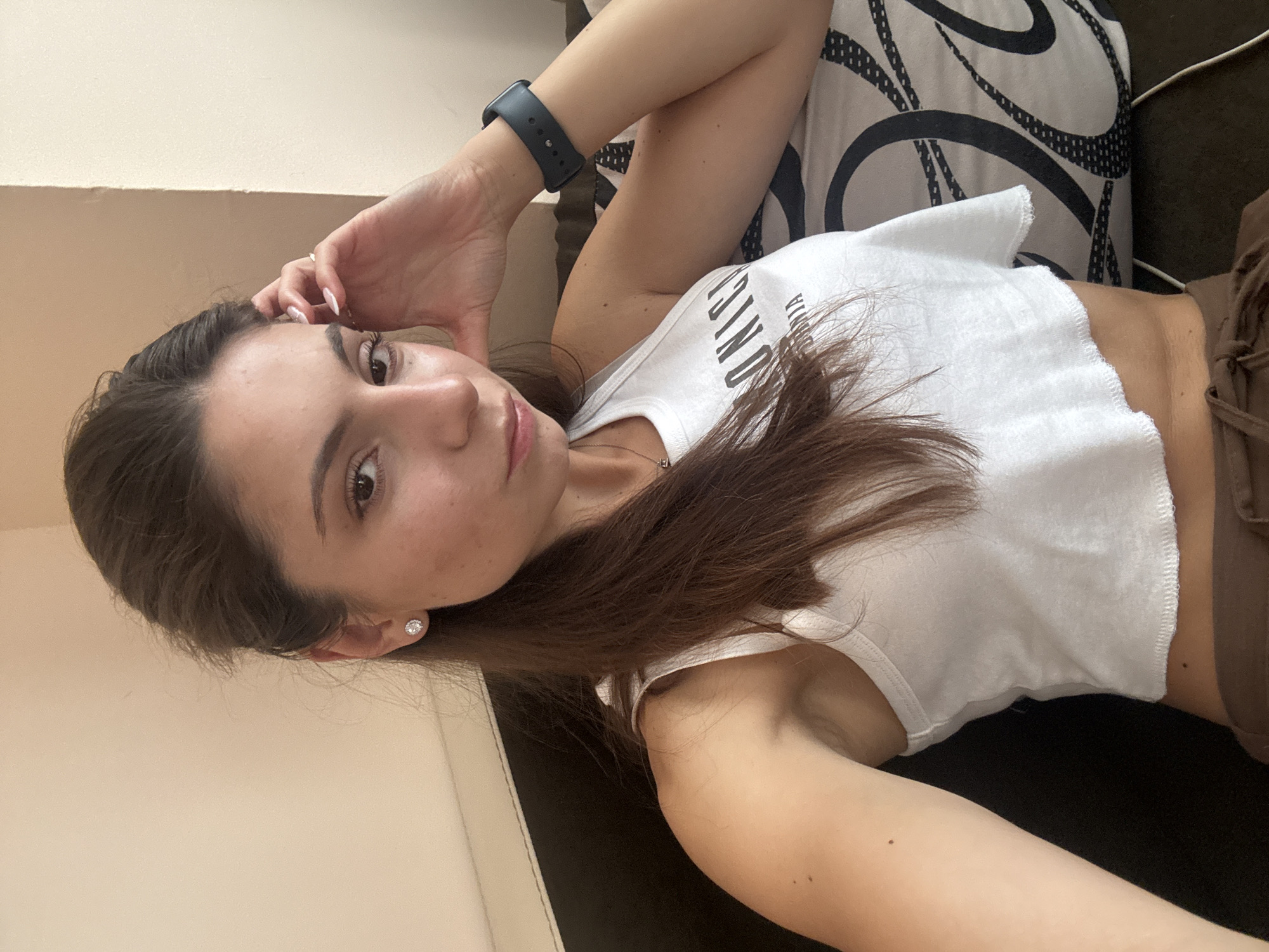 A woman is sitting indoors, possibly in an apartment in Varna, Bulgaria. She is positioned on a dark sofa with a patterned cushion. The woman has long, dark brown hair pulled back, a white tank top, and brown pants. She is looking towards the camera with her hand resting on her head. A dark smartwatch is visible on her left wrist. The lighting suggests it is daytime, with natural light coming from an unseen source.