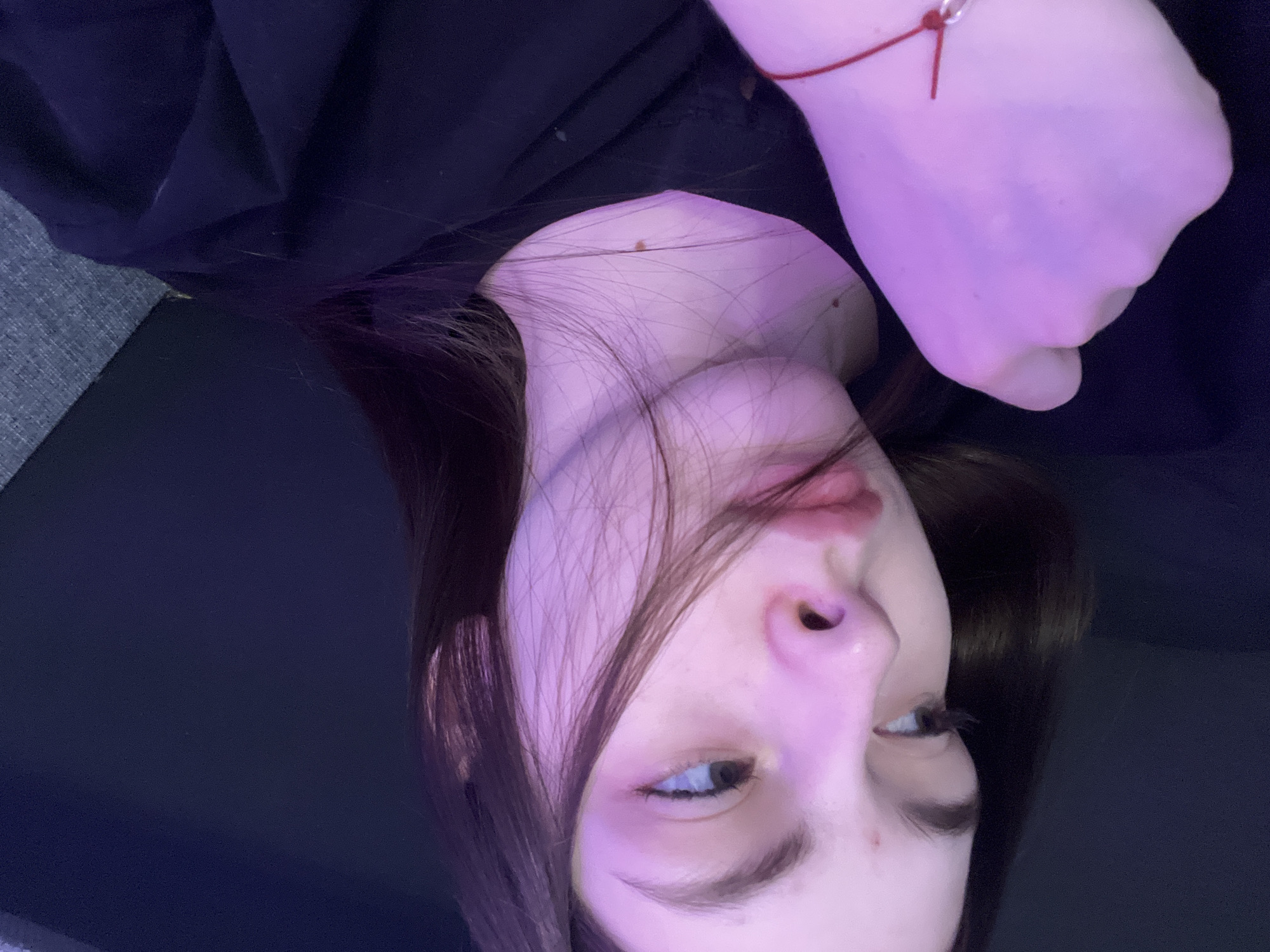 The image is a close-up selfie of a fair-skinned person with dark hair. The person is wearing a black shirt. The photo is taken from a low angle, looking up at the person's face, which is slightly tilted. The lighting is dim with a pinkish hue. A red string bracelet is visible on the person's wrist. The background is dark and indistinct. It is impossible to determine the location in Saint Petersburg, Russia from the provided image.