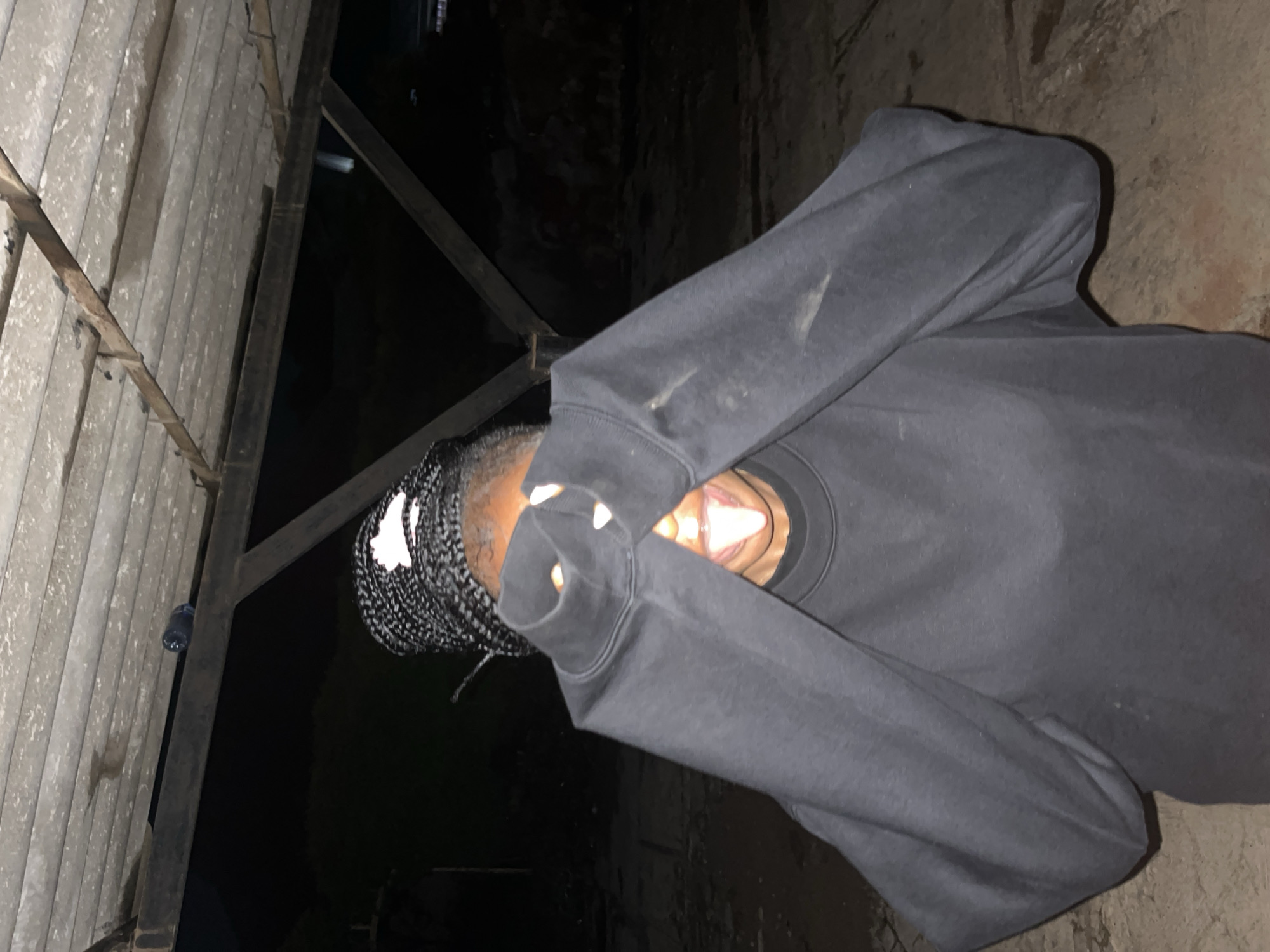 In Jos, Nigeria, a person with braided hair wearing a black hoodie is shown with their face covered by the hoodie's sleeves. The person's tongue is sticking out. The background consists of a dark, outdoor environment with a corrugated metal roof structure overhead. The ground appears to be dirt and debris. A plastic bottle is visible on a metal beam above and behind the person. The scene is dimly lit.