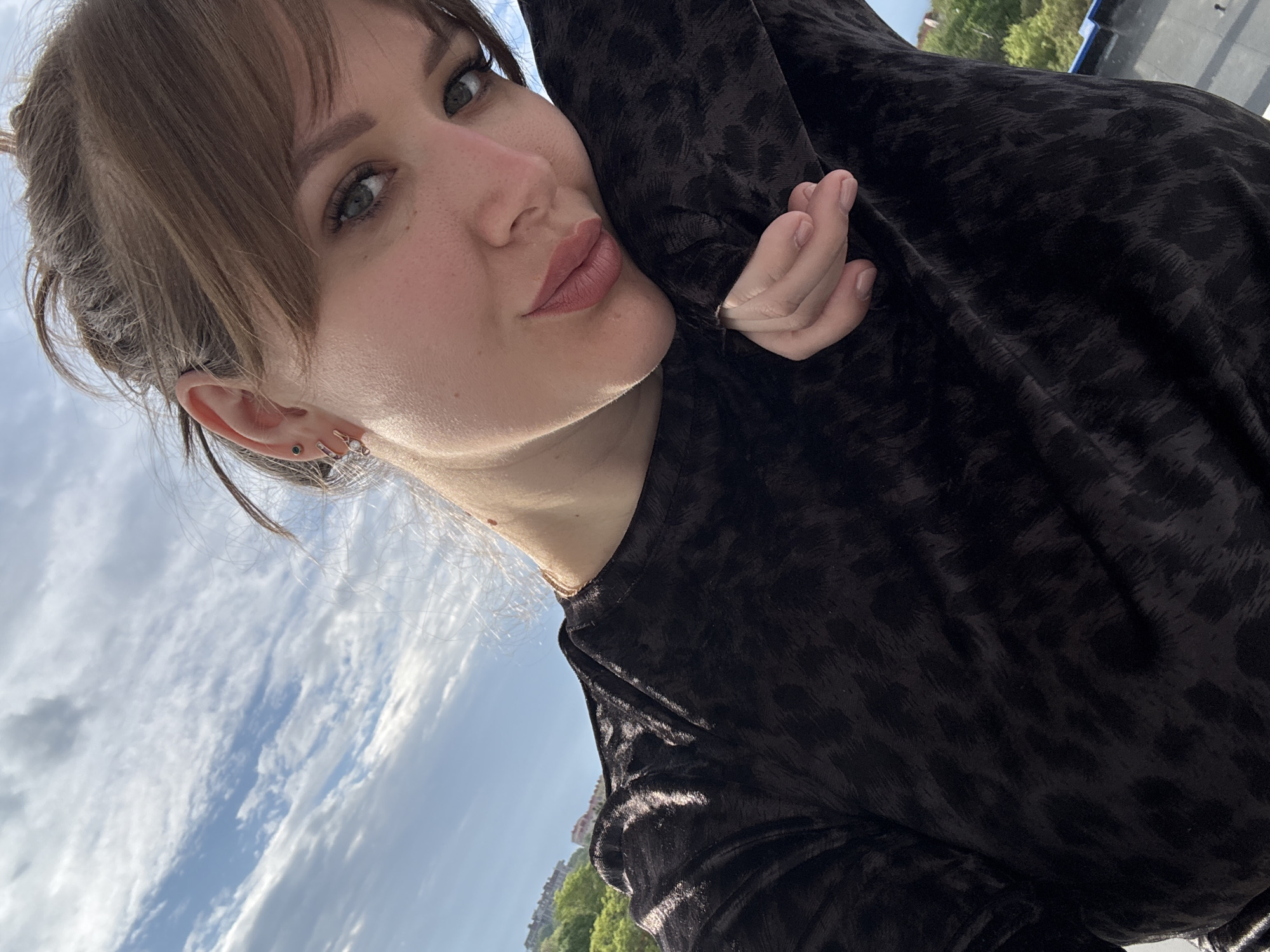 A woman poses for a close-up selfie against a backdrop of sky and urban scenery. The woman is fair-skinned, with brown hair styled with bangs, and is wearing a patterned brown top. She is holding her hand near her face and has earrings visible in her left ear. The background includes a blue sky with clouds, green trees, and buildings in the distance. The location is the city of Yevpatoriya, Ukraine.