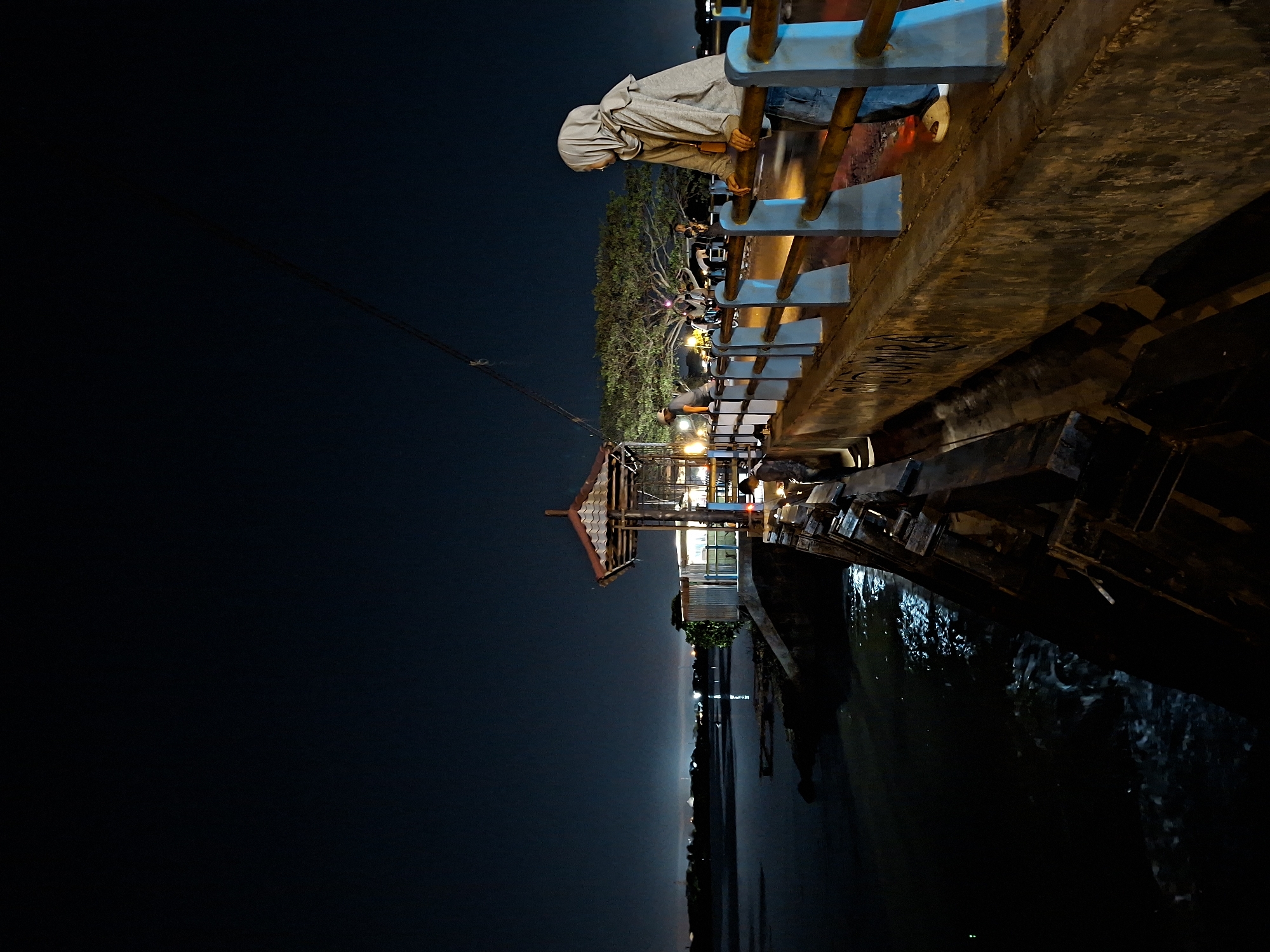 The image depicts a nighttime scene at the Tugu Bambu Runcing roundabout in Lhokseumawe, Indonesia. A woman wearing a light-colored hijab and a hooded jacket is leaning on a railing, looking out over the water. Her jeans and light-colored shoes are visible. The railing is made of wood with blue supporting posts. The background features a large tree illuminated by lights, with other people and vehicles visible near it. To the left, a structure with a red-tiled roof and a corrugated metal wall stands by the water's edge. The water appears dark and reflects some of the lights. The overall atmosphere is dimly lit, characteristic of a night scene.