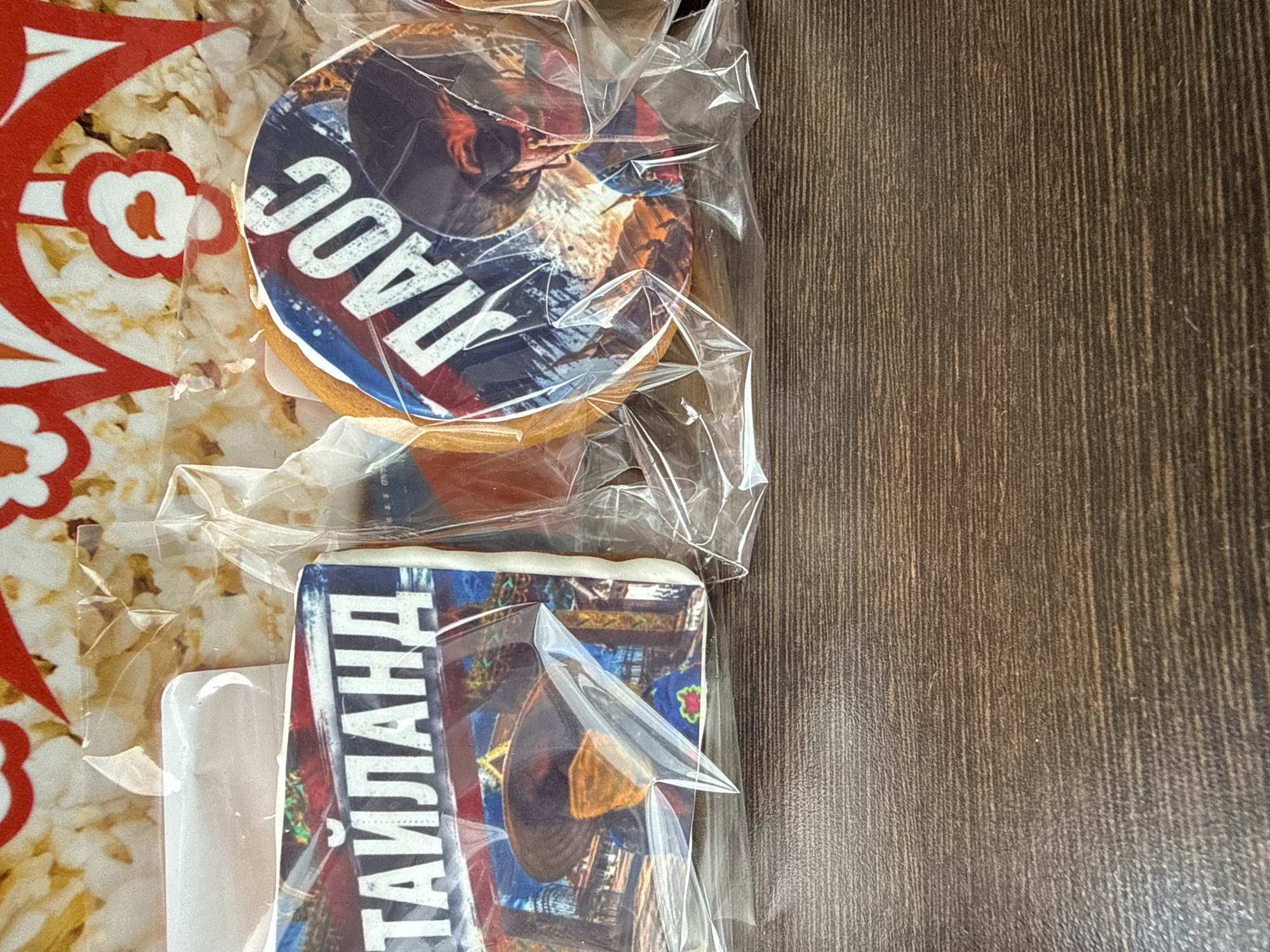Two individually wrapped cookies are displayed on a dark wooden surface. The cookie on the left is rectangular and features an image of a person wearing a wide-brimmed hat and traditional Thai attire, with the text "ТАЙЛАНД" (Thailand) printed in large, white, Cyrillic letters across the top. The cookie on the right is round and decorated with an image of a person wearing a cowboy hat and sunglasses, with the Cyrillic letters "ЛАДОС" printed in white. The cookies are enclosed in clear plastic packaging. The background is blurred, but appears to be a patterned surface, possibly related to popcorn packaging, with red and white decorative elements. There are no discernable landmarks or features that would indicate the city of Inkerman, Ukraine.