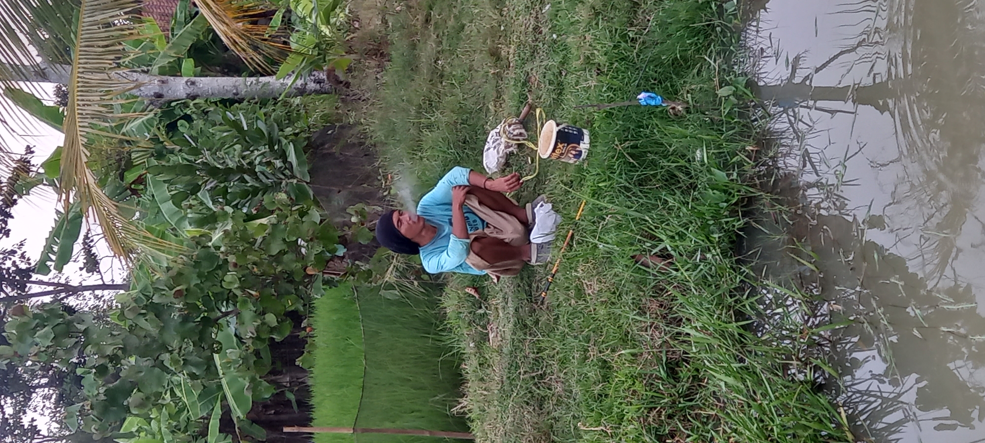 A man wearing a black head covering and a light blue long-sleeved shirt is squatting on a grassy area next to a body of water. He appears to be smoking and exhaling a cloud of smoke. In front of him, there are several objects including what appears to be fishing equipment, a white bucket with some markings, and a light-colored bag or bundle. The environment is lush and green with trees and foliage in the background, suggesting a rural or natural setting, possibly near rice paddies, which are characteristic of regions like Sidamulya, Indonesia. The water in the foreground reflects the surrounding vegetation.
