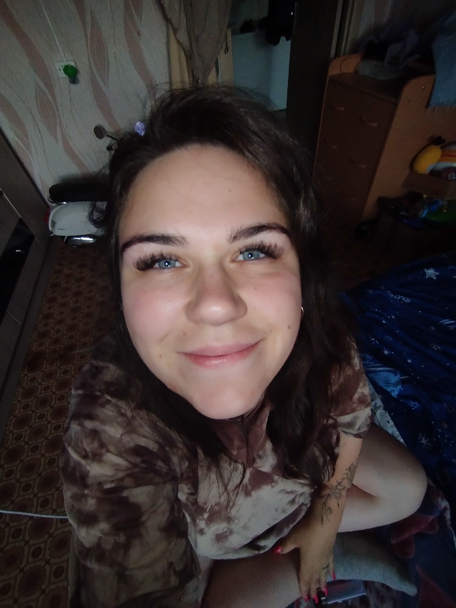 A young woman is featured in a close-up selfie, captured from a low angle, looking directly into the camera with a bright smile. She has dark hair, striking blue eyes with pronounced eyelashes, and a small hoop earring in her right ear. A faint purple clip or accessory is visible in her hair above her forehead. She is wearing a brown and beige tie-dye or patterned short-sleeved top. A detailed floral or leafy tattoo can be seen on her left forearm, and her fingernails are painted with bright pink polish.
The background indicates an indoor setting, likely a room in Chaikovsky, Russia. To the left, a wall is covered in light-colored wallpaper with wavy brown and beige patterns, and a green light or device is mounted on it. Below, a portion of a white and silver object, possibly a scooter or appliance, is visible on a dark brown patterned floor. To the right, a wooden dresser with multiple small handles is present, topped with a yellow plush toy and other items. A dark blue blanket adorned with white star patterns covers what appears to be a bed or couch. The lighting suggests an indoor environment, making the specific time of day unclear. No text is visible in the image. The woman's expression conveys a friendly and content mood.