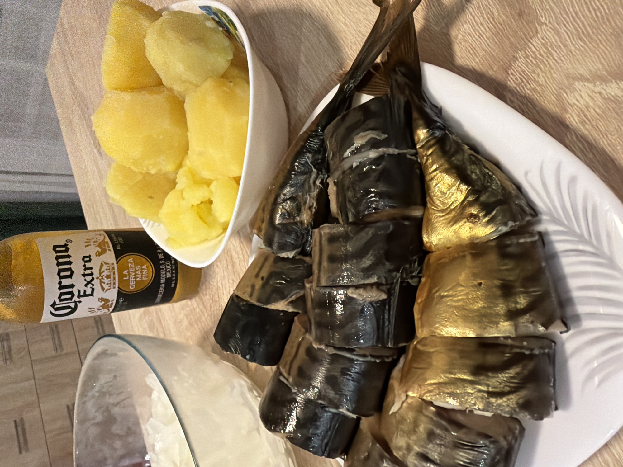 A close-up, eye-level shot captures a meal set on a wooden surface. In the foreground, a white plate holds several pieces of smoked fish, cut into sections, arranged in two overlapping rows. The fish exhibits a dark, almost black exterior and a golden-brown underside. To the right of the fish, a white bowl contains boiled potatoes, cut into chunks. Behind the fish and potatoes, a clear glass bowl contains what appears to be chopped onions or a similar white food item. A brown glass bottle of Corona Extra beer stands upright to the left of the bowl of potatoes. The background shows a wall with horizontal wooden paneling. The lighting is bright and direct. The scene appears to be indoors, likely in a kitchen or dining area, with no discernible elements suggesting a specific location within Moscow, Russia. There are no people present in the image, and no actions or interactions are depicted.